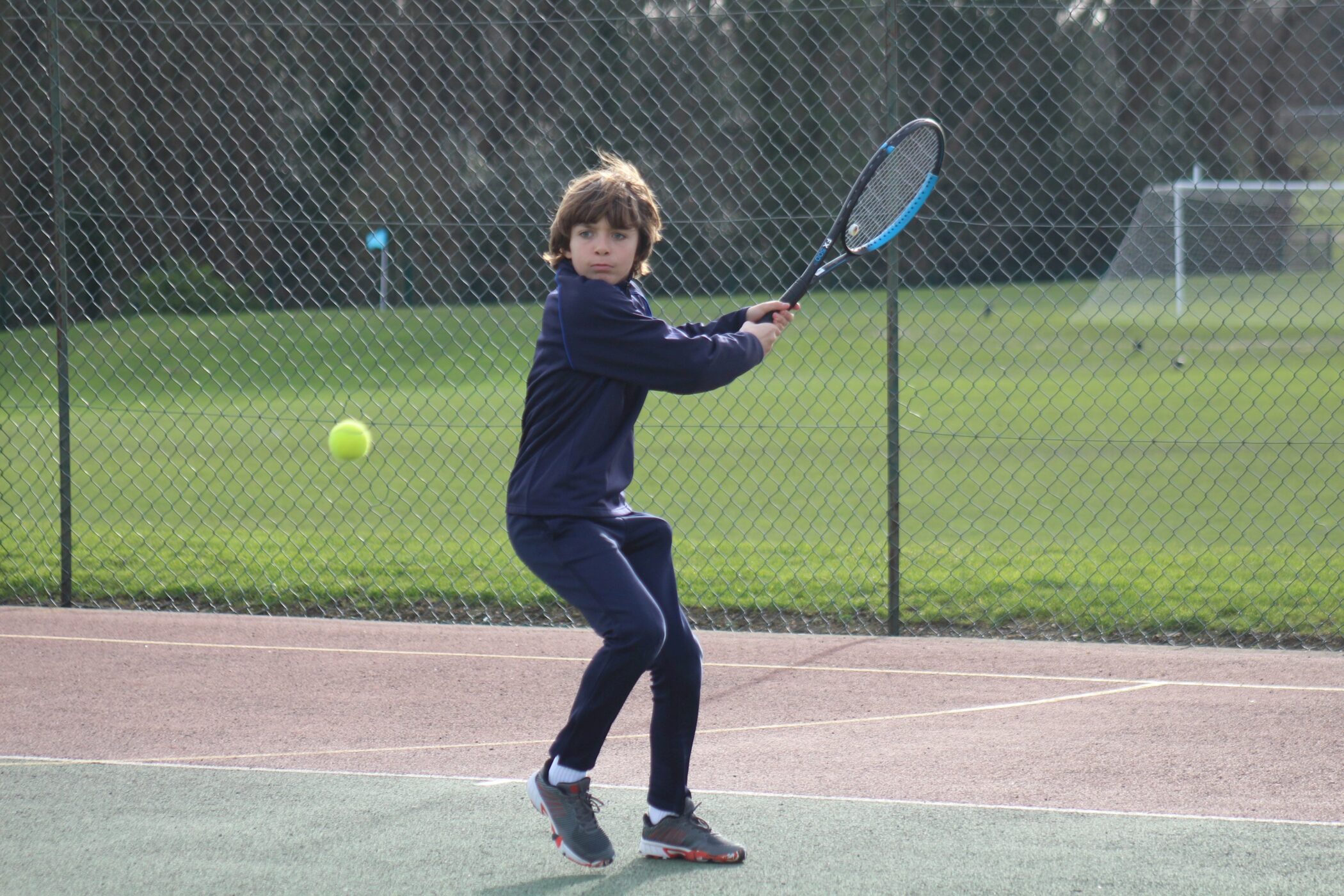A young boy in a navy tracksuit prepares to hit a tennis ball with a racquet on an outdoor court, with a chain-link fence and grass field in the background.