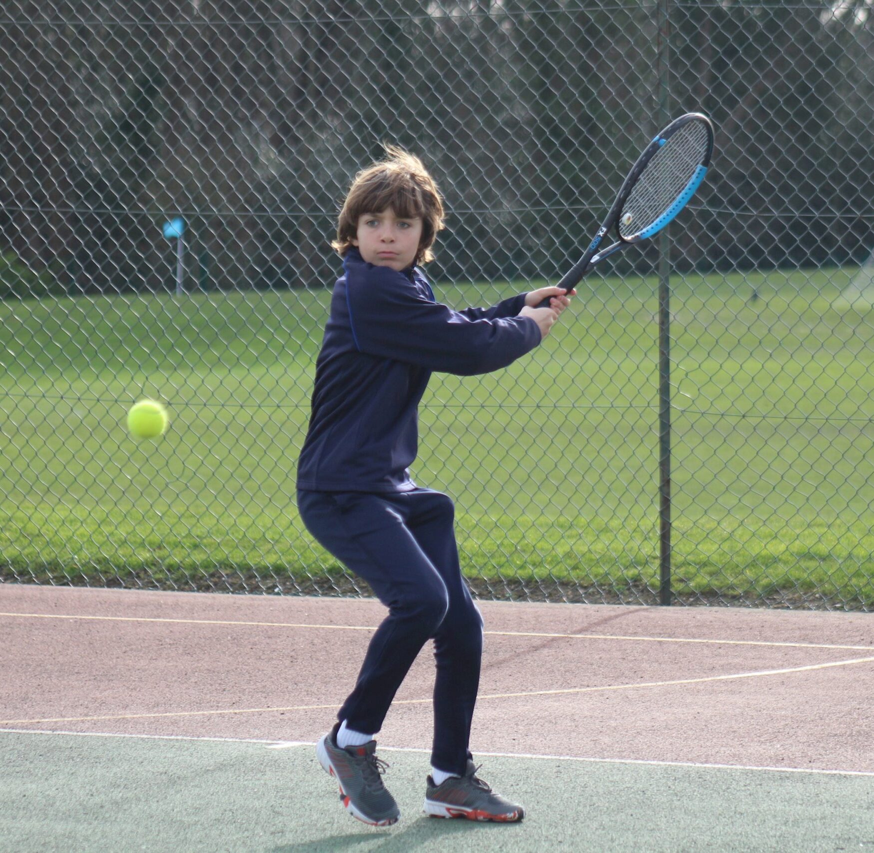A young boy in a navy tracksuit prepares to hit a tennis ball with a racquet on an outdoor court, with a chain-link fence and grass field in the background.