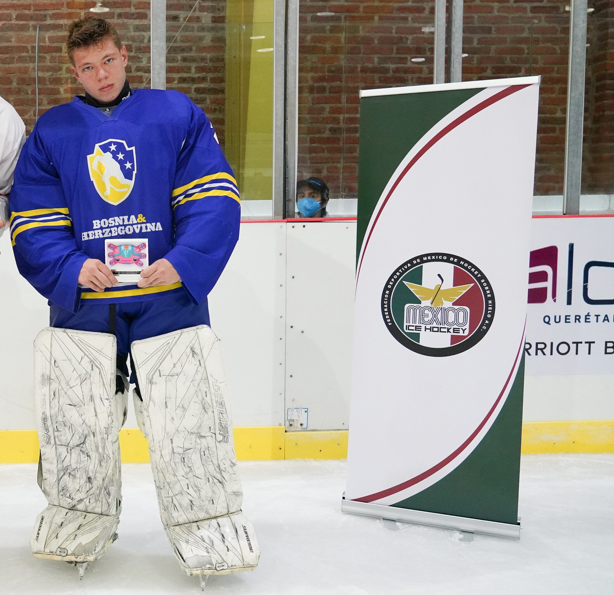 A hockey player in a blue Bosnia & Herzegovina jersey and white goalie pads stands on the ice holding an award, with a person in a mask visible behind the rink glass.