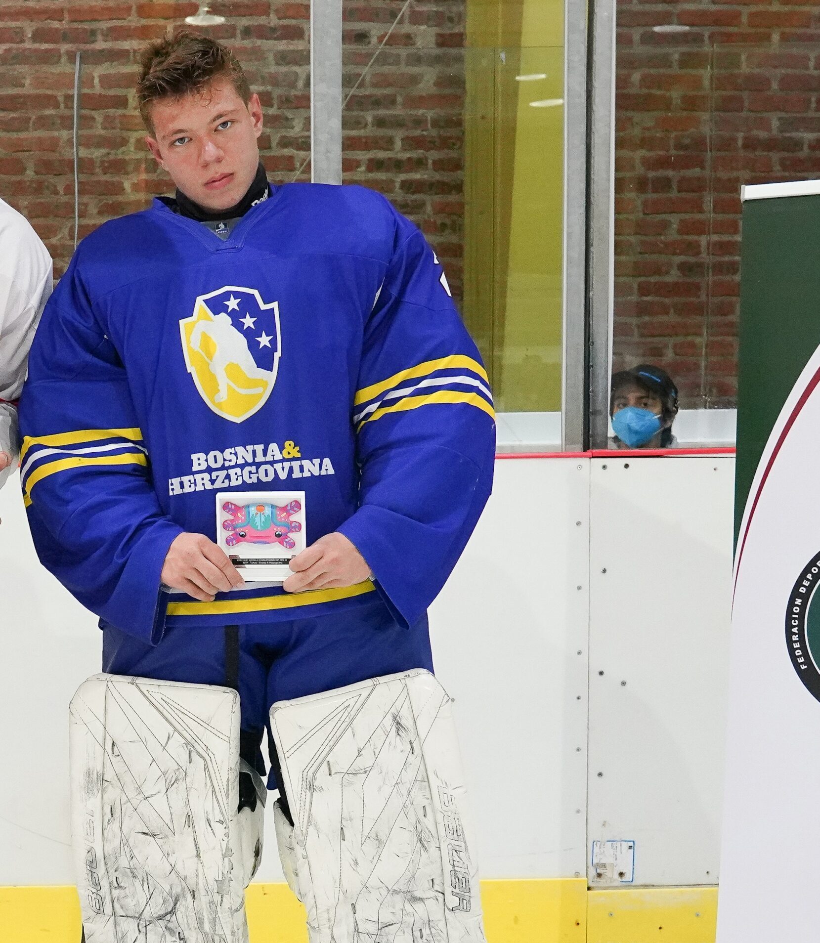 A hockey player in a blue Bosnia & Herzegovina jersey and white goalie pads stands on the ice holding an award, with a person in a mask visible behind the rink glass.