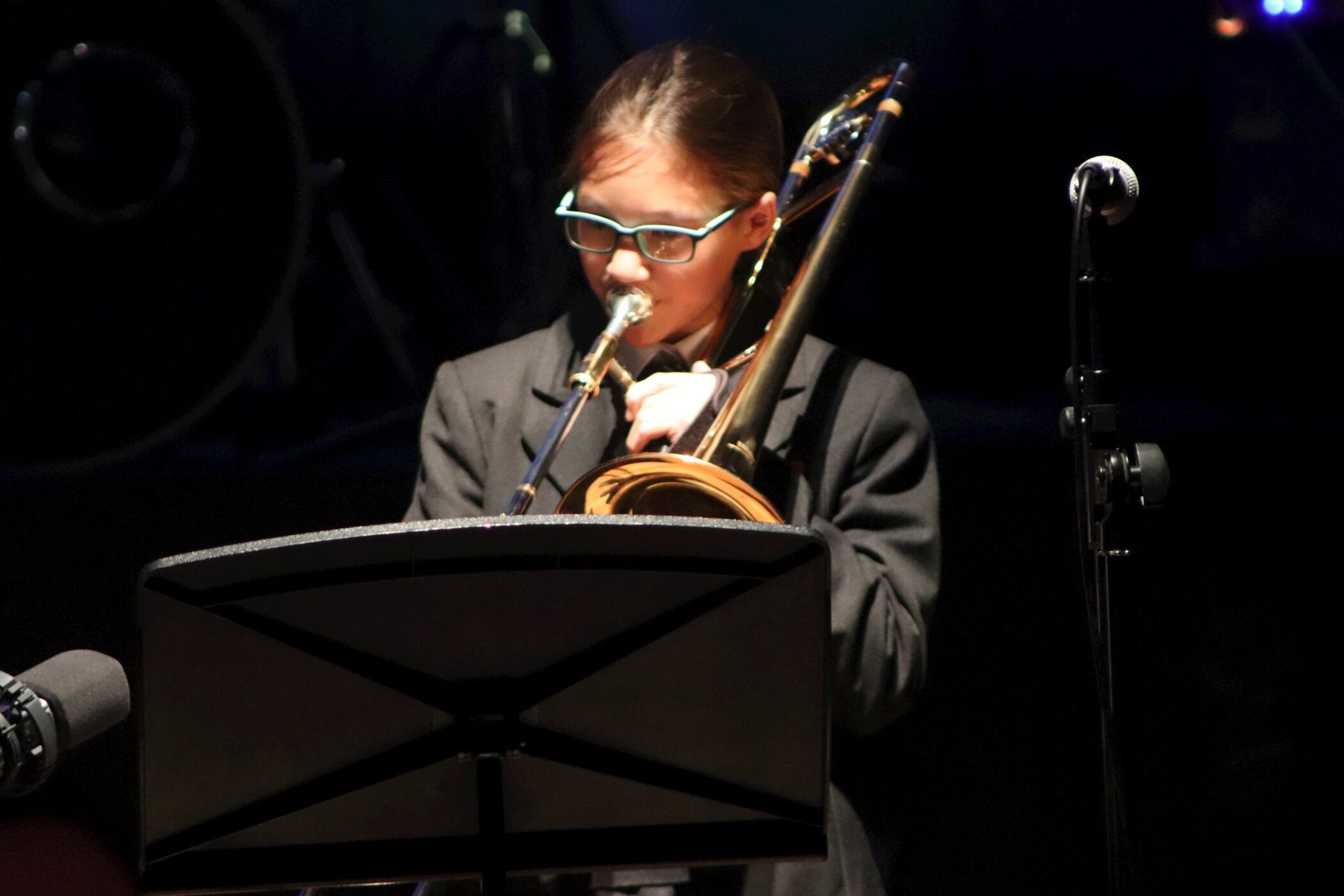 A young person wearing glasses plays a brass instrument, likely a trombone, while reading sheet music on a stand. The individual is dressed in a dark suit, and the background is dimly lit.