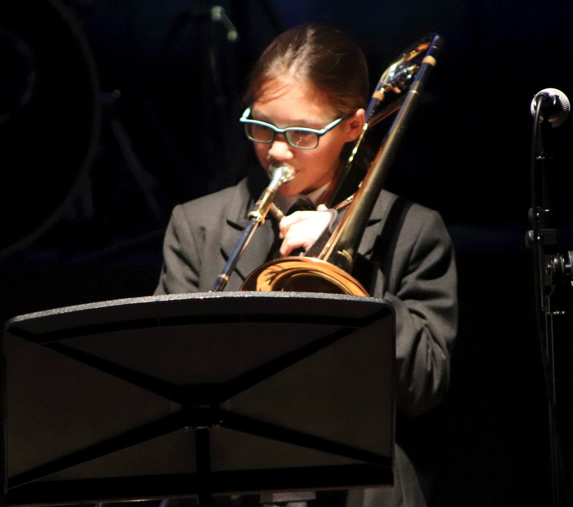 A young person wearing glasses plays a brass instrument, likely a trombone, while reading sheet music on a stand. The individual is dressed in a dark suit, and the background is dimly lit.