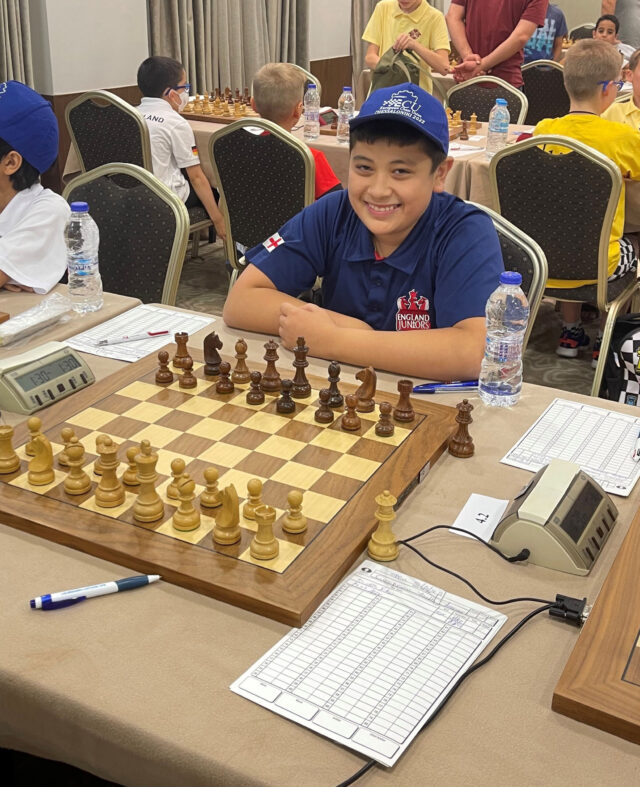 A boy in a navy cap and England team shirt smiles at a chessboard, ready to play in a tournament. Chess clocks, score sheets, and water bottles are on the table. Other children are seated and standing in the background.