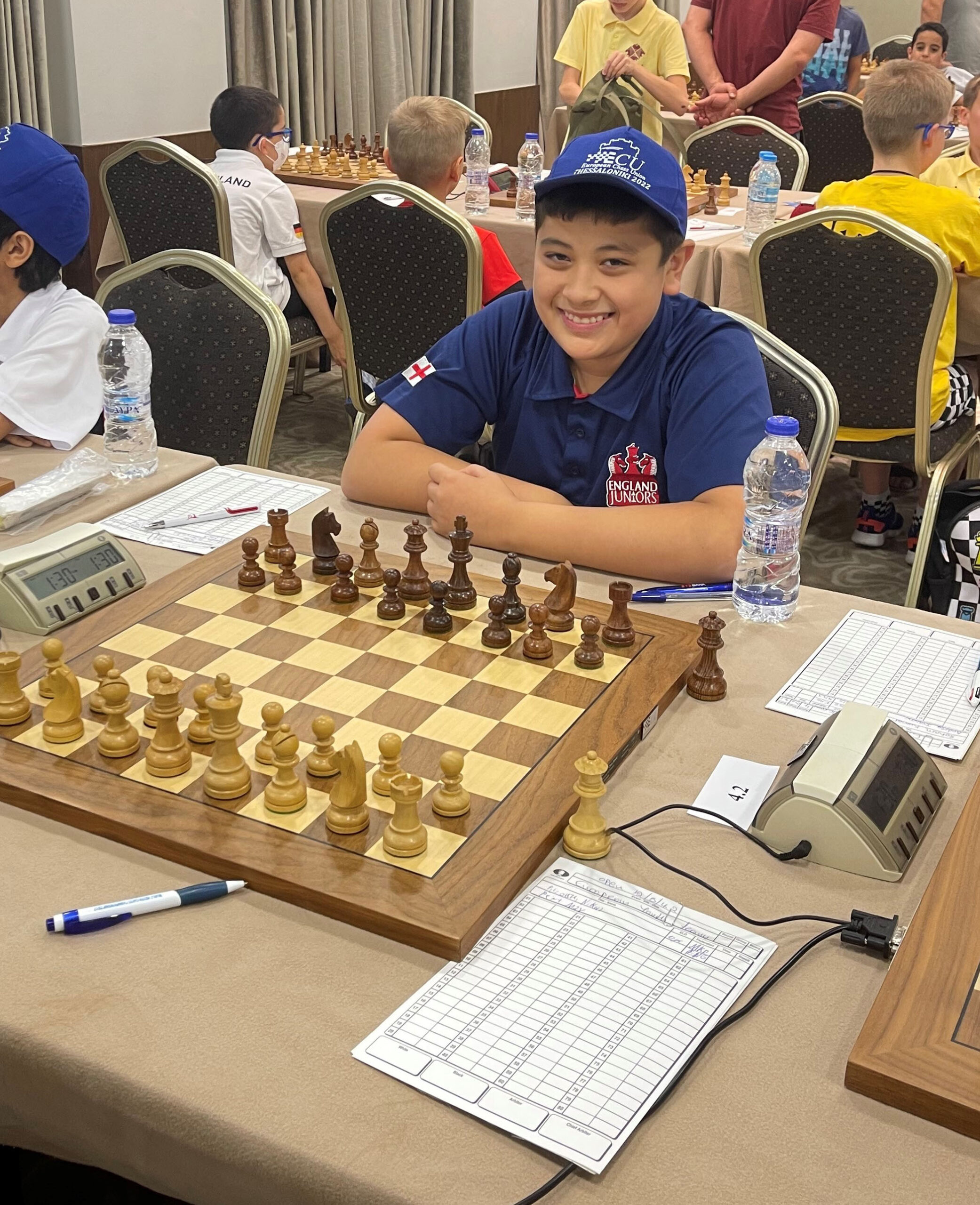 A boy in a navy cap and England team shirt smiles at a chessboard, ready to play in a tournament. Chess clocks, score sheets, and water bottles are on the table. Other children are seated and standing in the background.