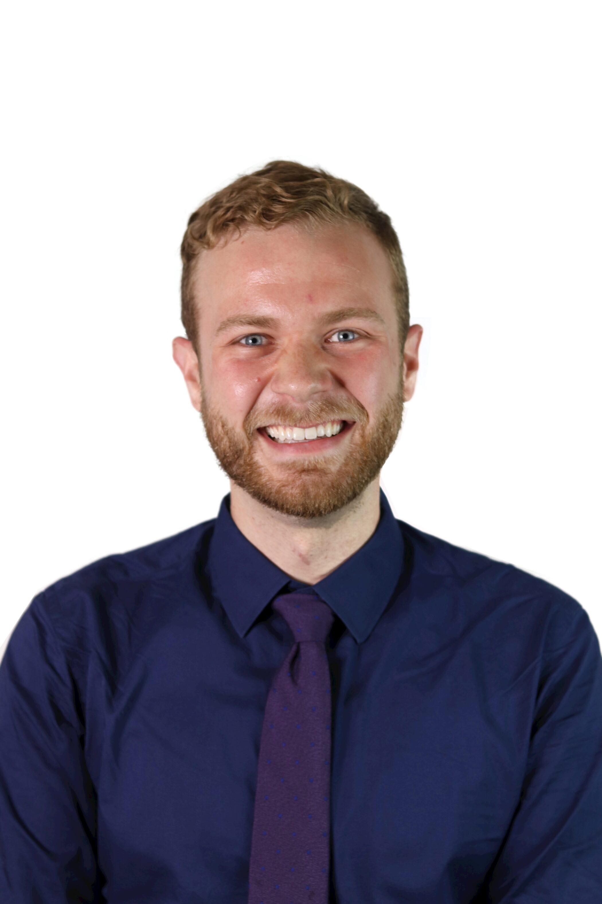 A man with short curly hair and a beard, wearing a dark blue shirt and matching tie, smiles at the camera against a plain white background.