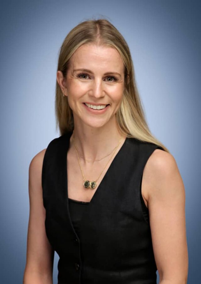 A woman with long blonde hair smiles at the camera. She is wearing a sleeveless black top, gold earrings, and a gold necklace with green stones. The background is a smooth gradient of blue.