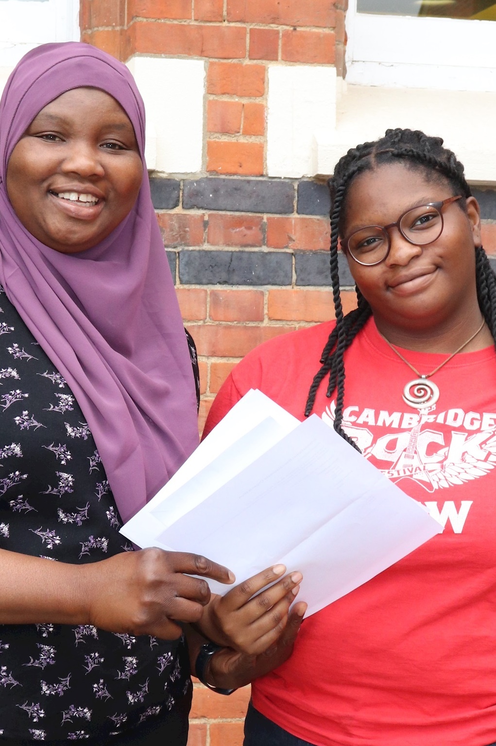 Two people smiling and holding papers, standing in front of a brick wall. One wears a purple hijab and floral top, the other wears glasses and a red Cambridge T-shirt.