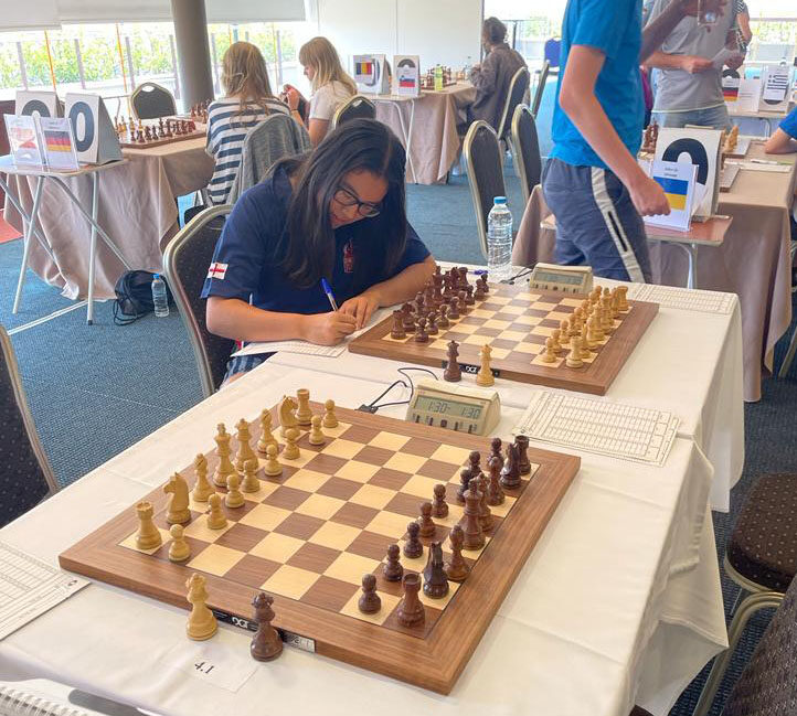 A young chess player with long dark hair writes on a scoresheet at a chess tournament. Two chessboards and a clock are on the table, and other players are visible in the background.