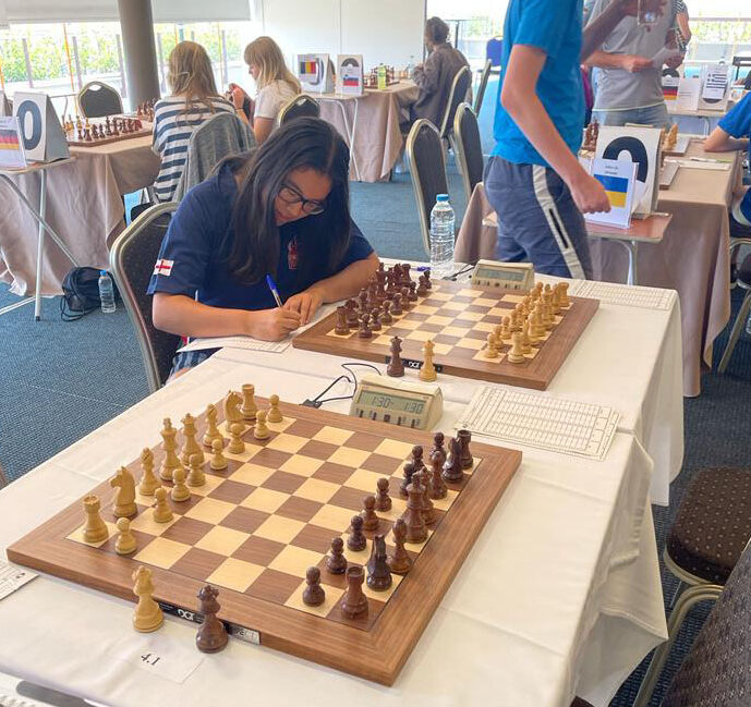 A chess tournament setting with multiple boards; a young person with long hair and glasses records a move at one table, while others stand and walk in the background. Pieces are set for the start of a match.