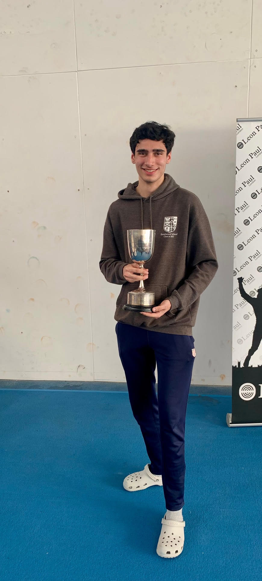 A young man in a dark hoodie and navy trousers stands on blue flooring, smiling and holding a silver trophy. He is wearing white Crocs. A banner with text and a silhouette is partially visible behind him.