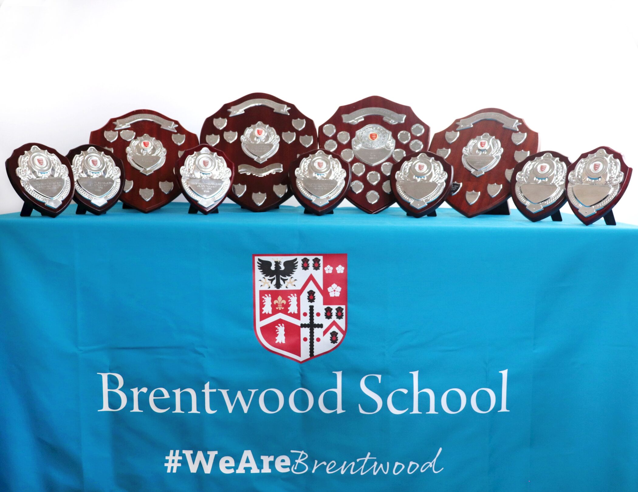 A row of silver and wooden shields and trophies displayed on a blue tablecloth with the Brentwood School crest and the hashtag #WeAreBrentwood.