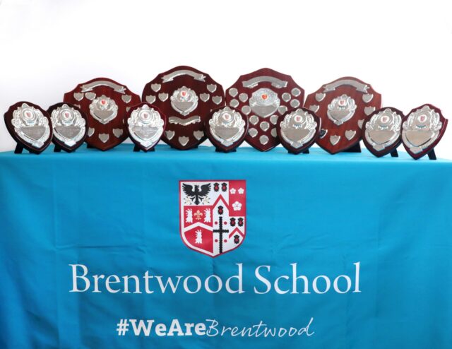 A row of silver and wooden shields and trophies displayed on a blue tablecloth with the Brentwood School crest and the hashtag #WeAreBrentwood.