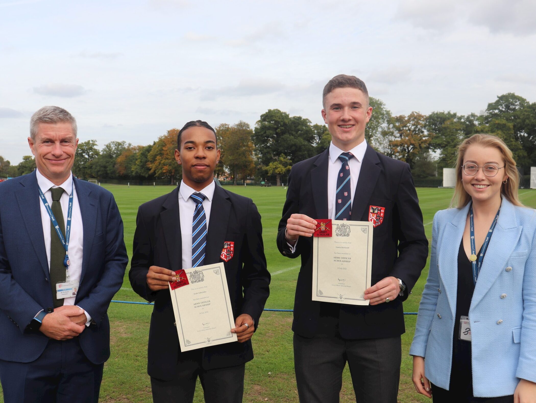 Four people stand outdoors on grass, two young men in blazers holding certificates, flanked by a man in a suit and a woman in a blue blazer, all smiling at the camera with trees in the background.