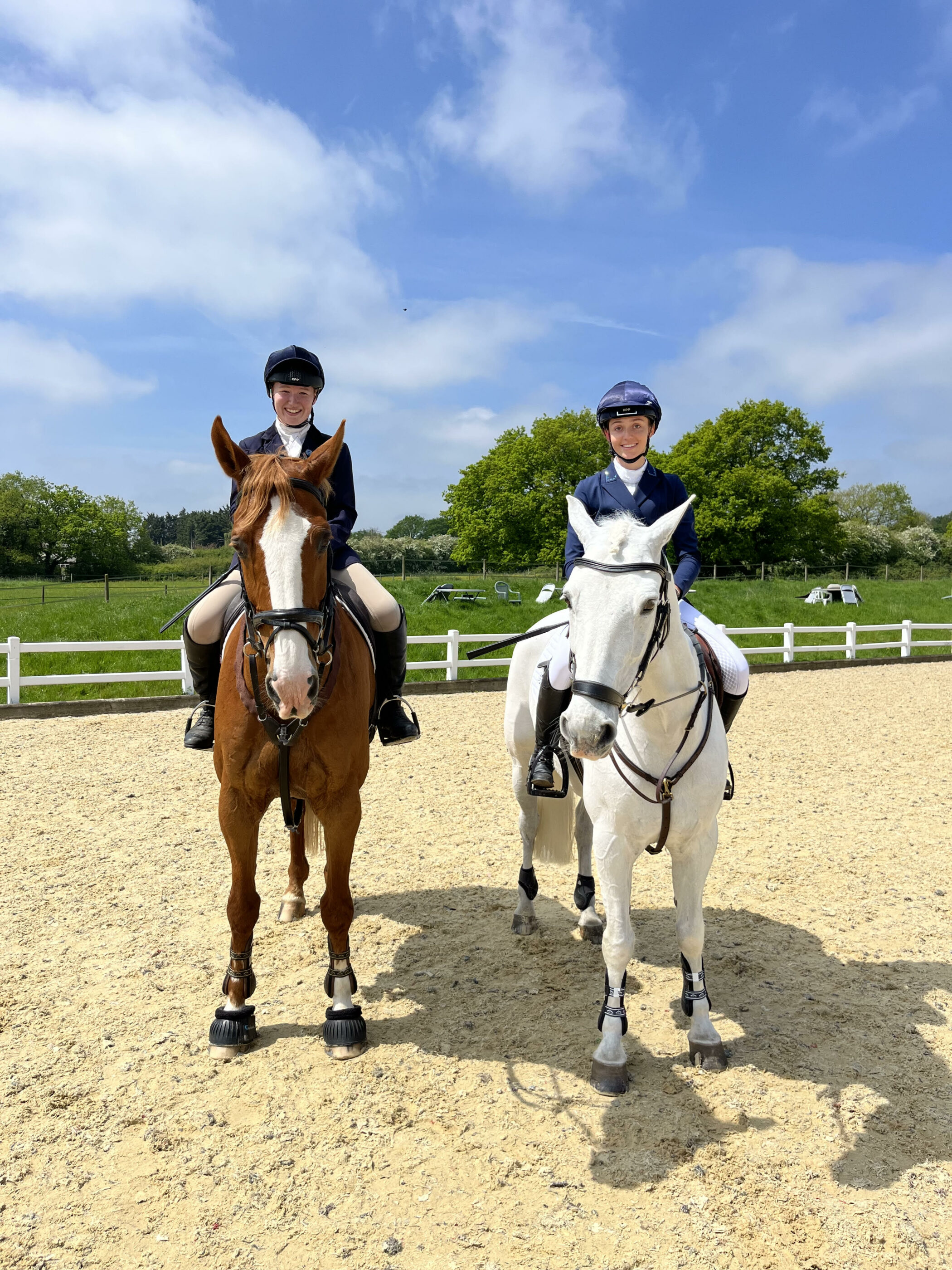 Two people in riding kit sit on horses, one brown and one white, in a sunny outdoor arena with a sandy surface, white fencing, and green trees in the background under a partly cloudy sky.