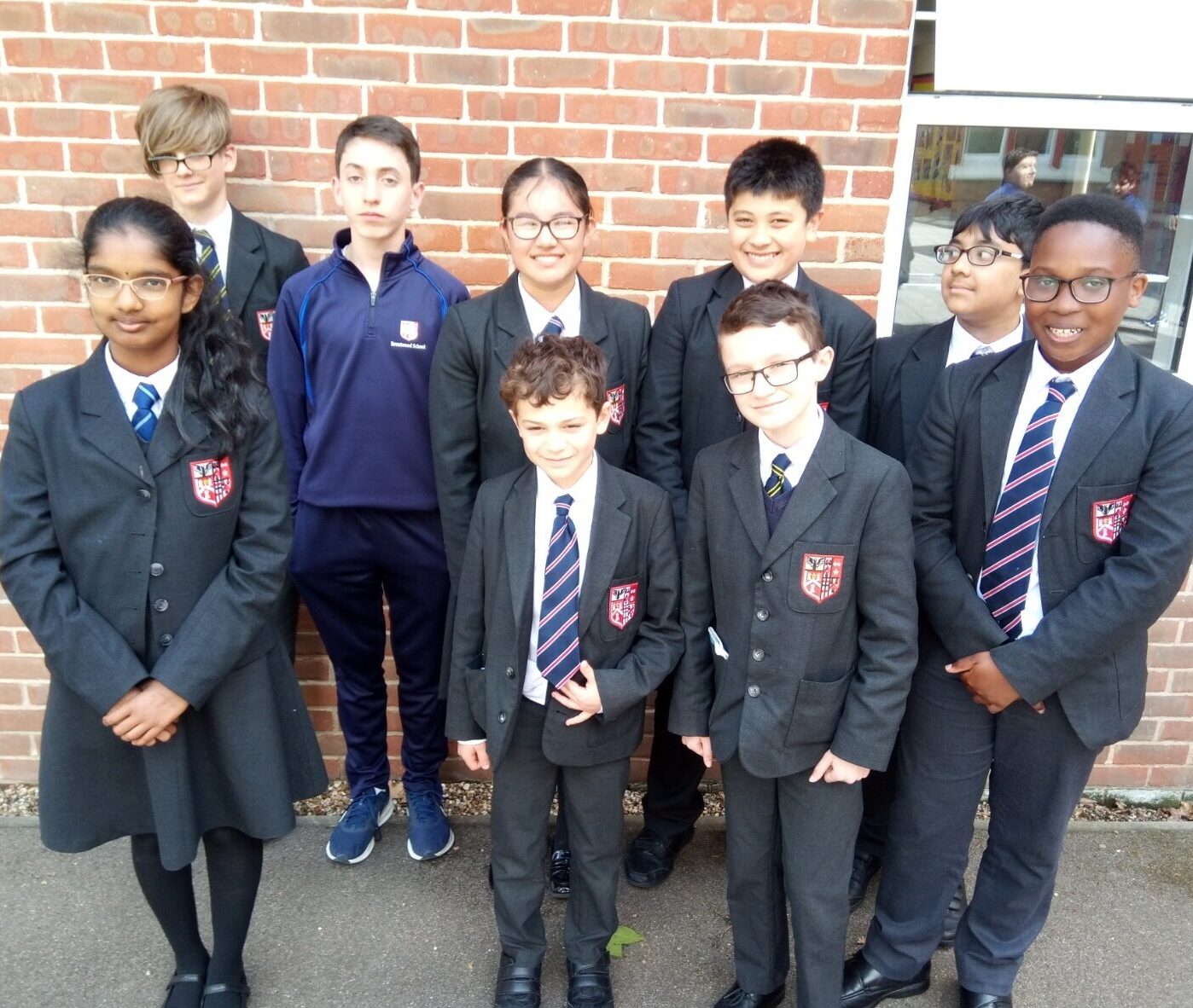 A group of nine schoolchildren, wearing dark blazers, ties, and glasses, stand together smiling in front of a brick wall. One child wears a navy tracksuit. They appear to be part of a school group or team.