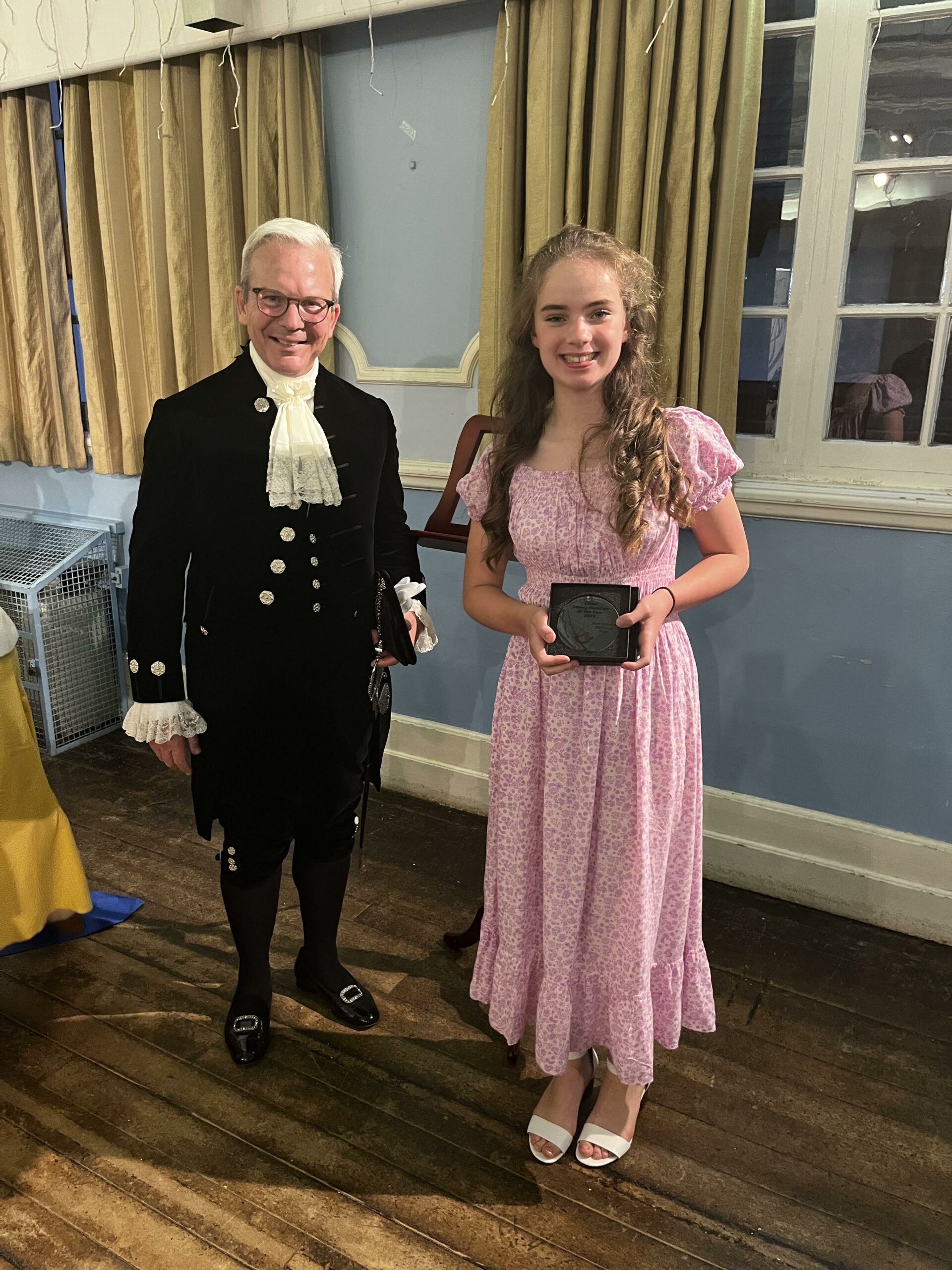 A man in formal black attire with a white cravat stands next to a smiling young woman in a pink floral dress and white heels, holding a small black box, in a room with wooden floors and gold curtains.