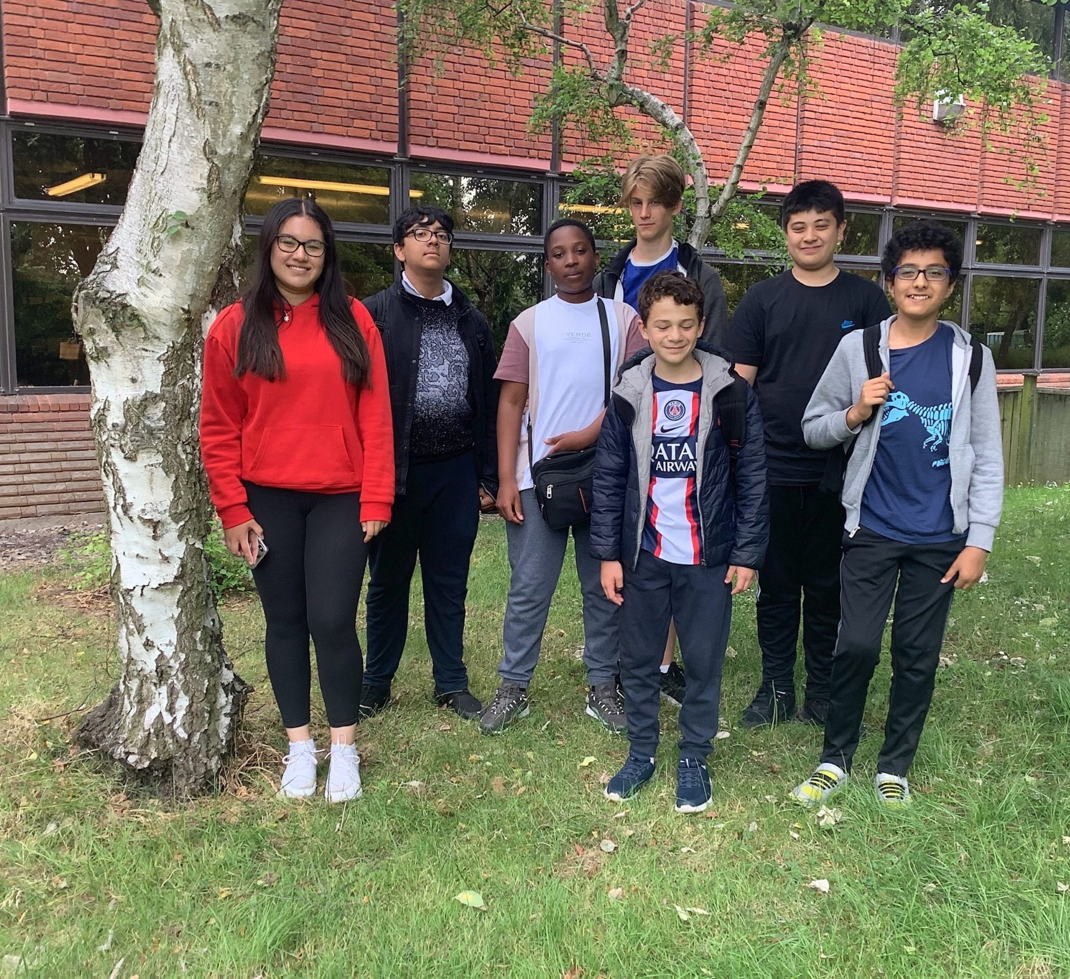 A group of seven teenagers stands on grass near a tree, posing for a photo in front of a building with large windows and a red brick wall. They are dressed in casual clothing and smiling at the camera.