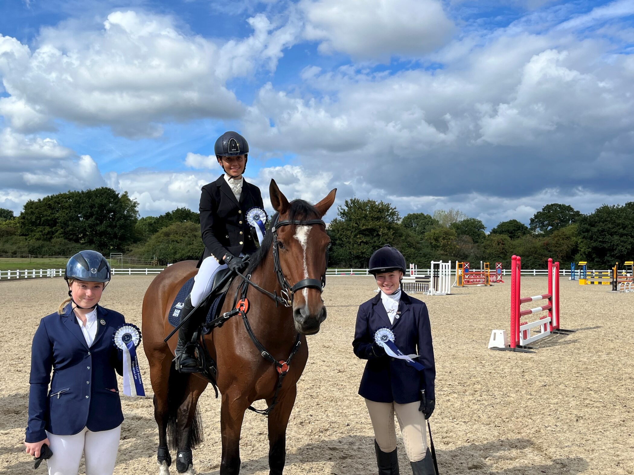 Three equestrians in riding kit pose at an outdoor arena. One is mounted on a brown horse, while the other two stand on either side, all holding blue and white rosettes. The sky is partly cloudy with trees in the background.
