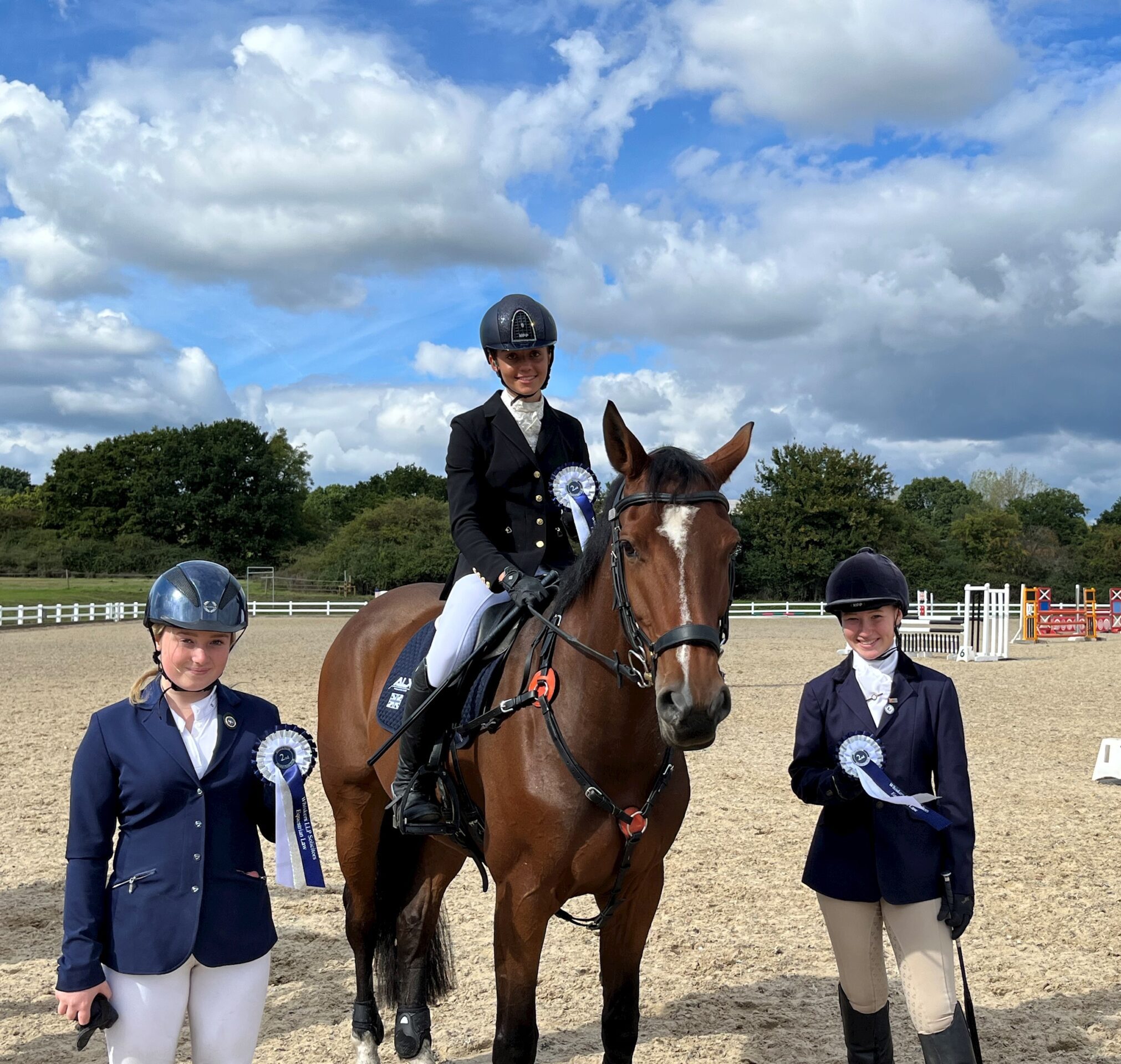 Three equestrians in riding kit pose at an outdoor arena. One is mounted on a brown horse, while the other two stand on either side, all holding blue and white rosettes. The sky is partly cloudy with trees in the background.