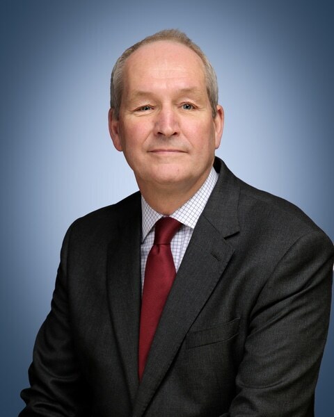A middle-aged man in a dark suit, white chequered shirt, and red tie sits against a blue gradient background, looking at the camera with a neutral expression—an image that could easily represent a Brentwood School Bursar.