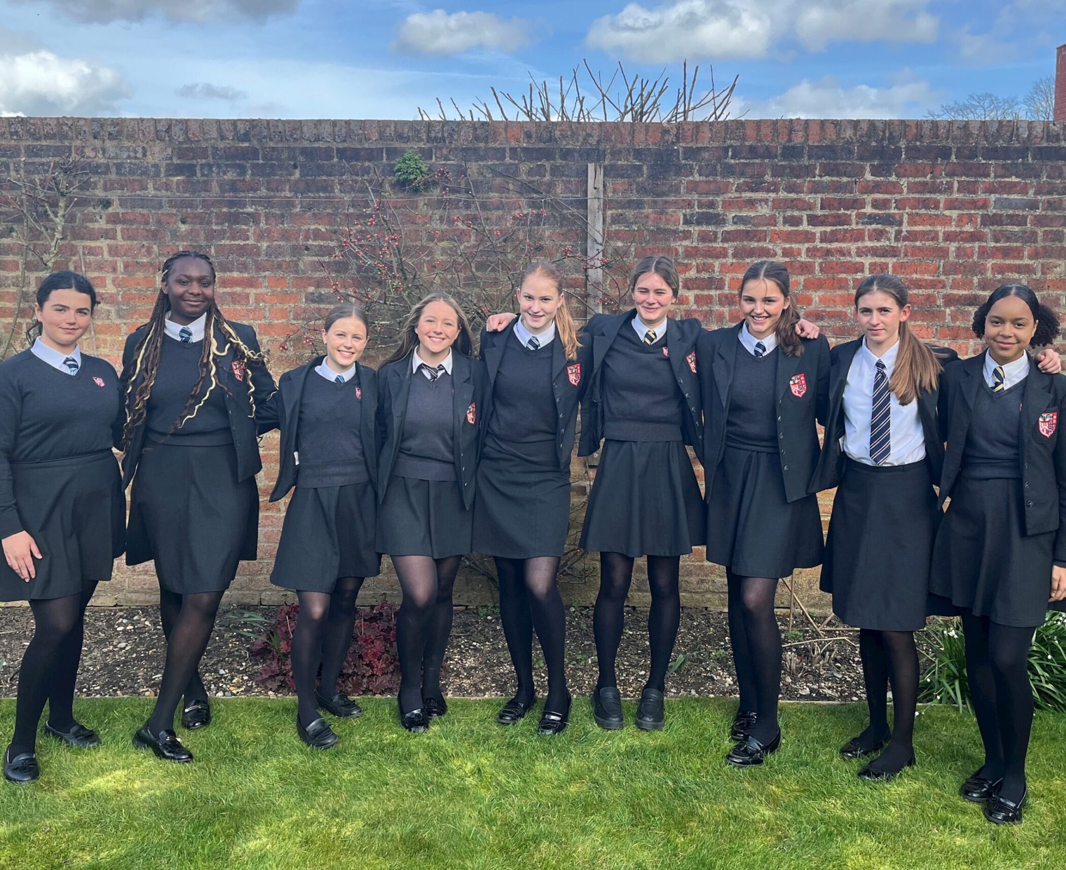 Eight girls in black school uniforms stand side by side, smiling, in front of a brick wall and garden. Most wear blazers and ties, and the weather appears clear and sunny.