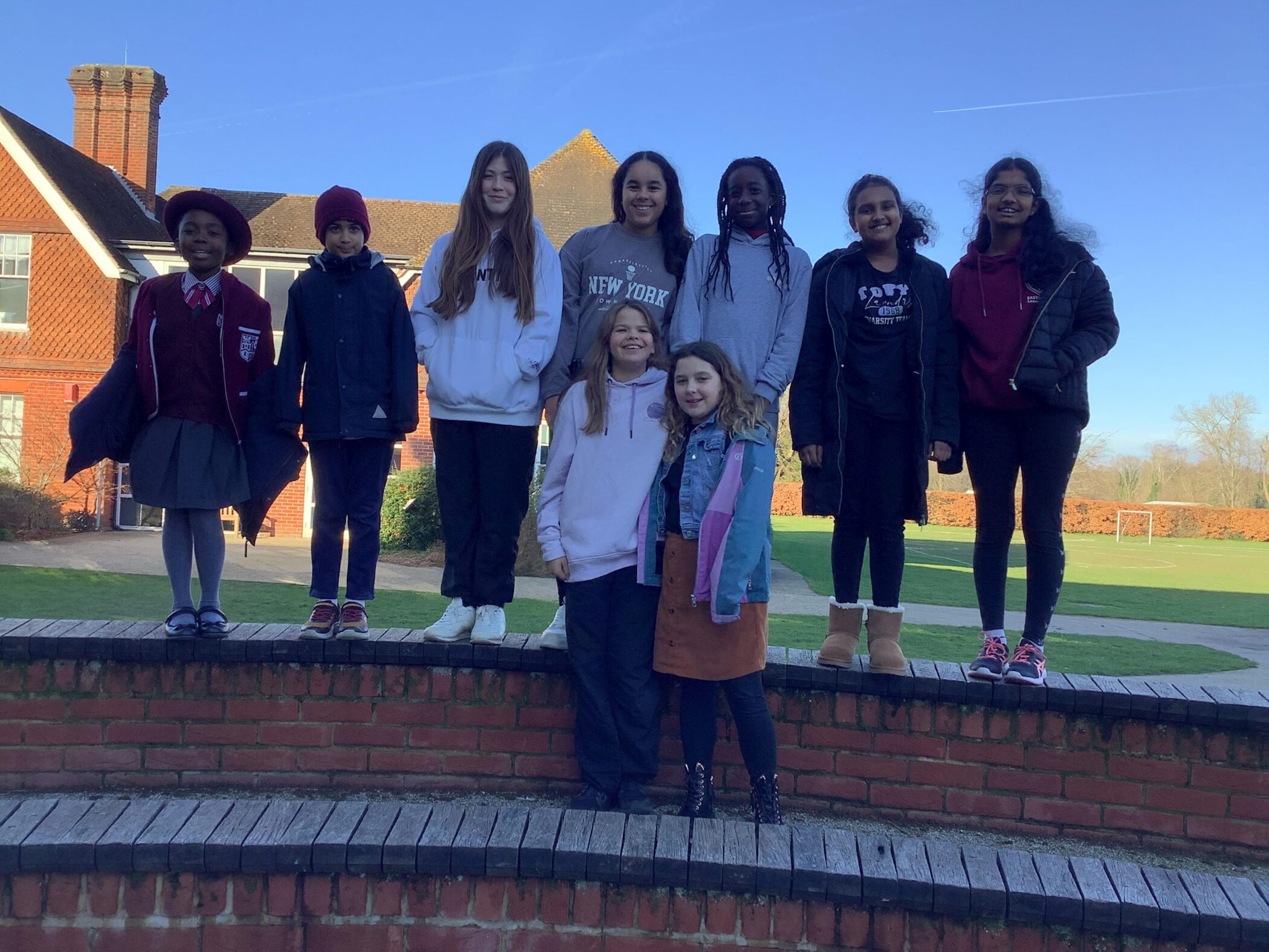 A group of nine children, eight standing on a low brick wall and two standing in front, pose outdoors on a sunny day with a school building and green lawn in the background.
