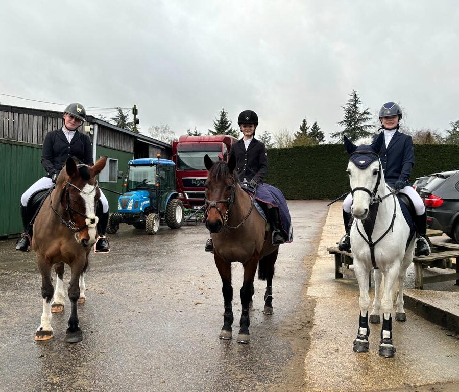 Three people in riding attire sit on horses in an outdoor stable area with vehicles, including a blue tractor and a red lorry, parked behind them. The ground is wet and the sky is overcast.