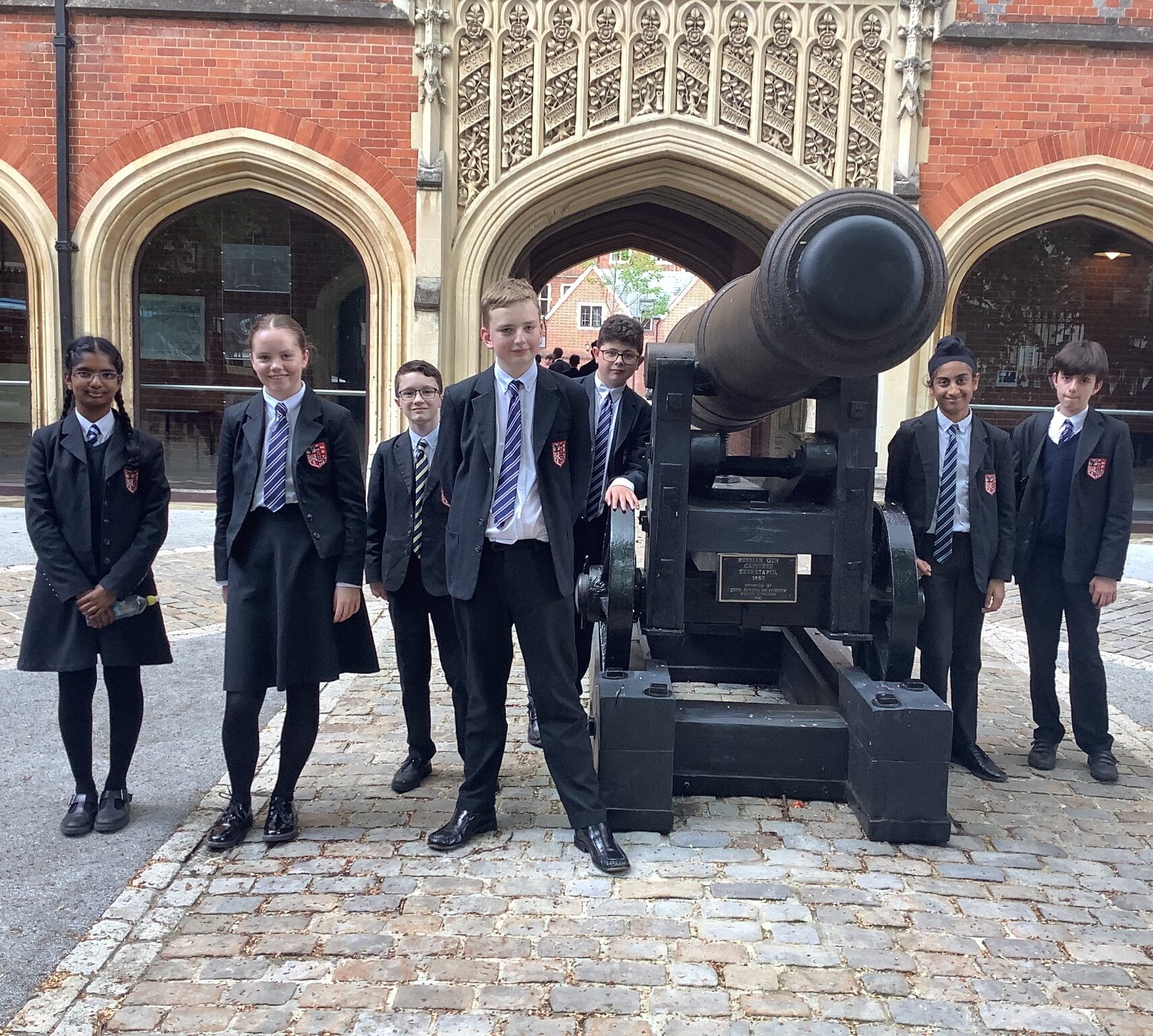A group of seven students in school uniforms pose in front of a large black cannon, set on cobblestones, with a historic brick building and arched doorways in the background.