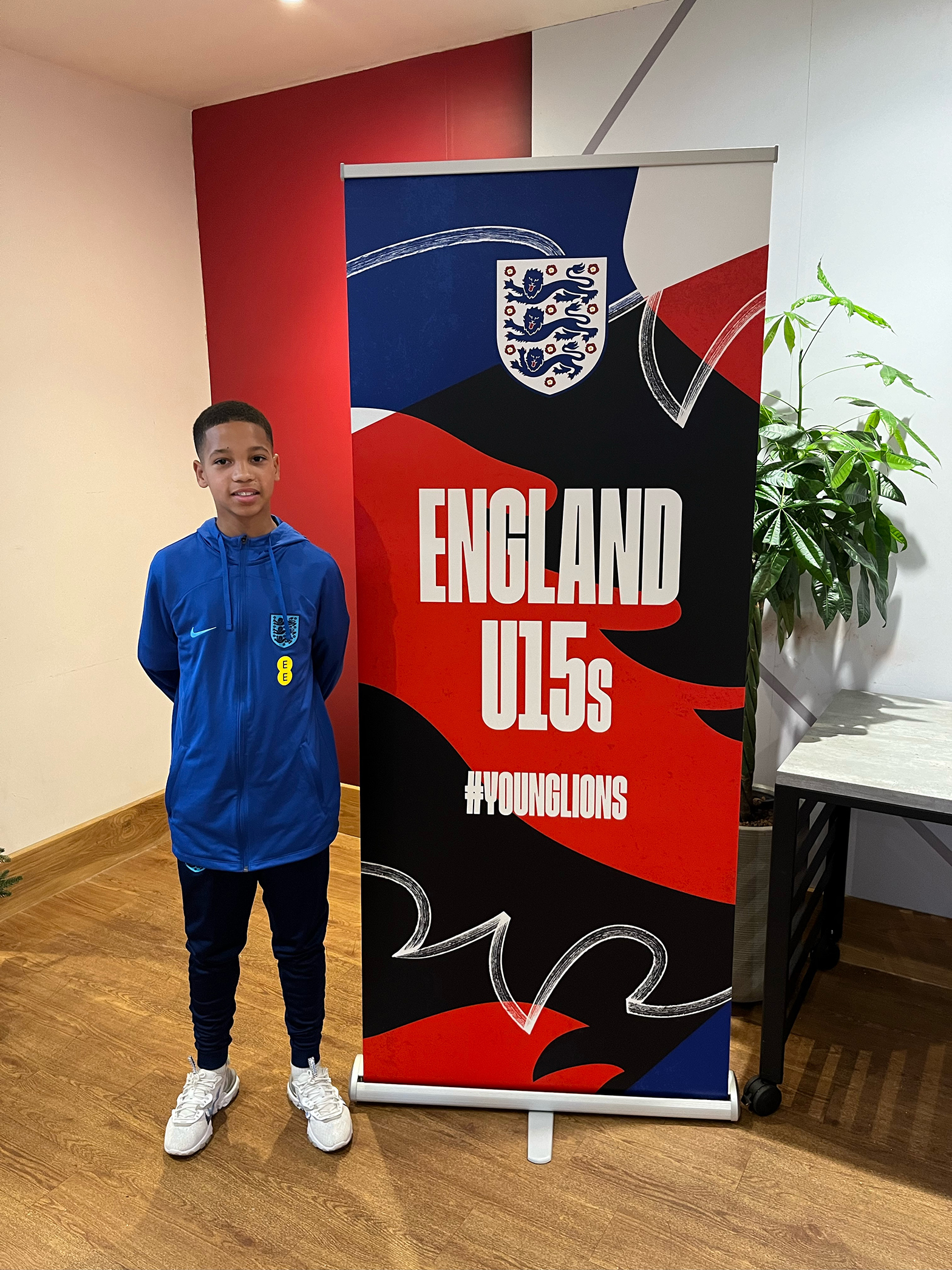 A young boy in an England football tracksuit stands next to a colourful England U15s “#YOUNGLIONS” banner, inside a room with wooden flooring and a green plant in the background.