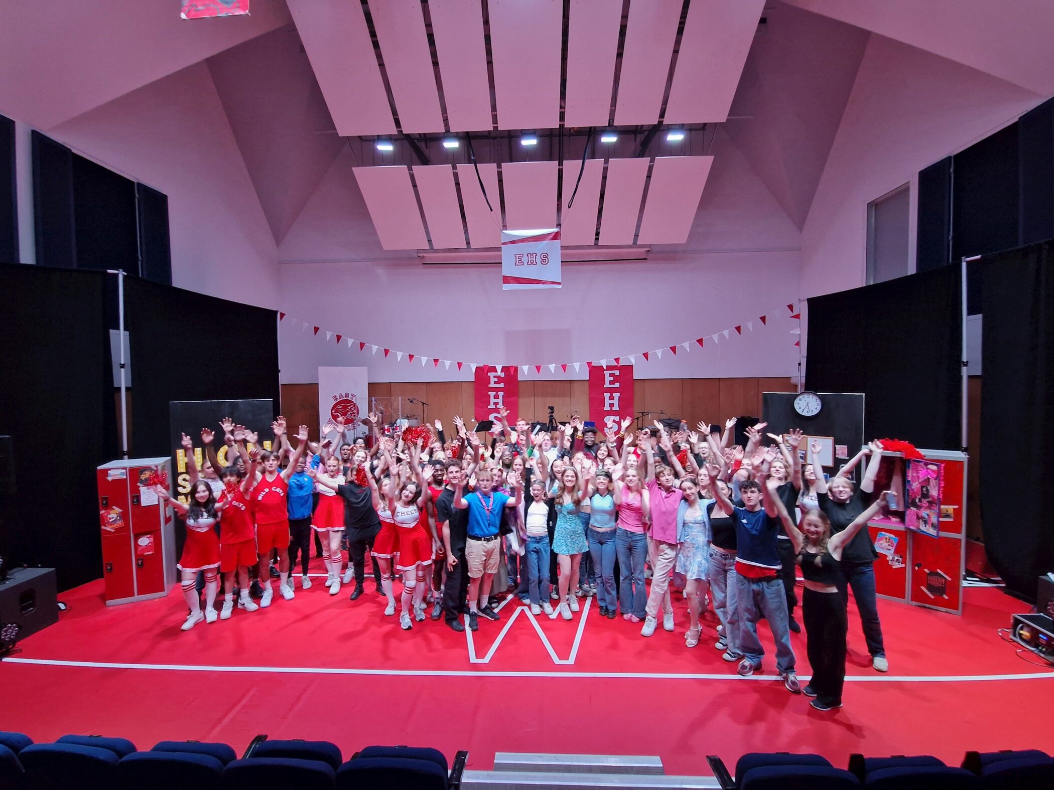 A large group of people, many in red and white cheerleader or sports outfits, stand on a red stage with hands raised, under white ceiling panels and High School Musical-themed decorations.