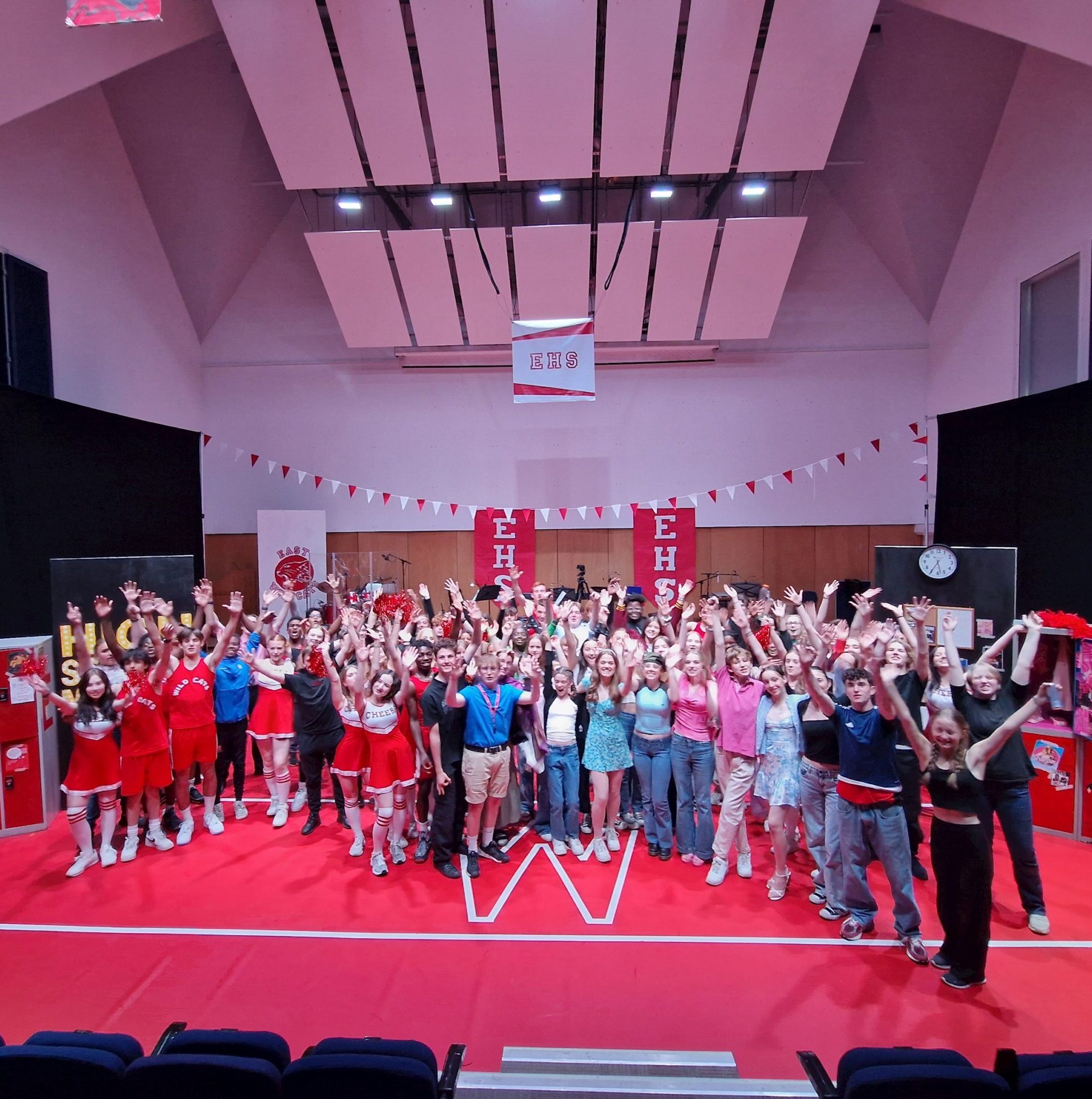 A large group of people, many in red and white cheerleader or sports outfits, stand on a red stage with hands raised, under white ceiling panels and High School Musical-themed decorations.