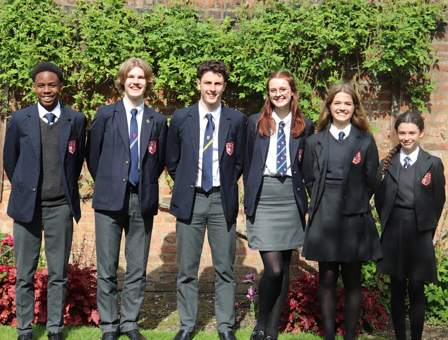 Six students in school uniforms stand in a row outside, smiling at the camera. They are in front of a brick wall covered with green plants and stand on grass with red flowers at their feet.