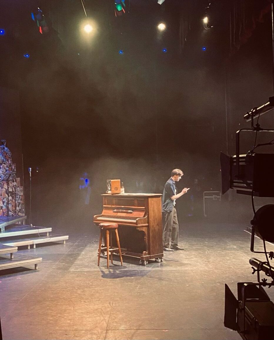 A person stands on a dimly lit stage near an upright piano and stool, appearing to hold papers or a script, with stage lights and equipment visible in the background.
