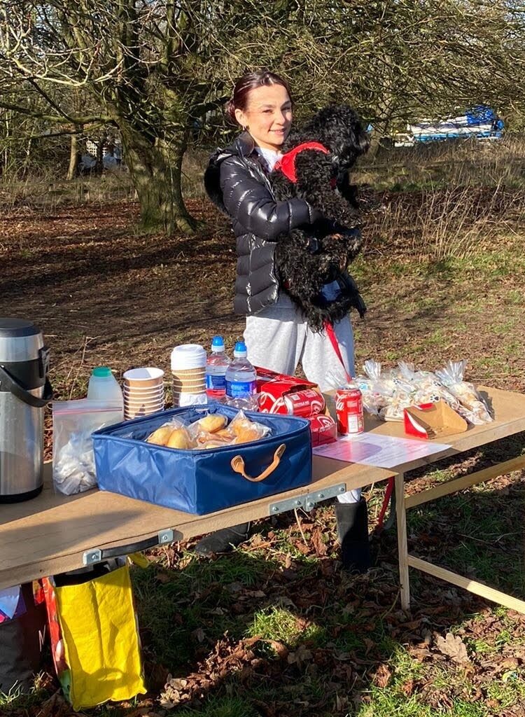 A woman in a black jacket smiles whilst holding a black dog in her arms, standing behind an outdoor table with drinks, snacks, and baked goods on a sunny day in a park surrounded by trees.