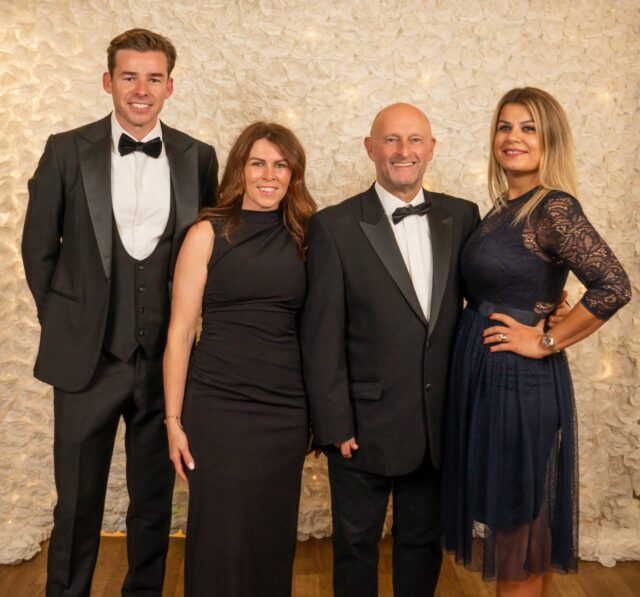 Four people from the Parents' Association stand in front of a light-coloured floral backdrop. The two men wear black dinner jackets, while the two women wear dark dresses, all smiling and posing for the photo.