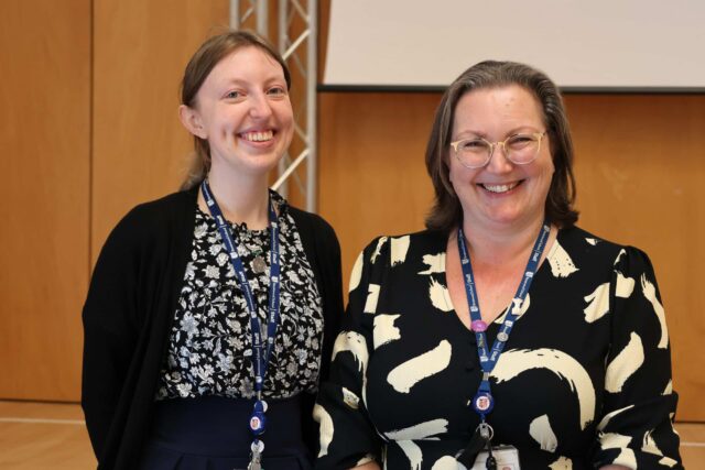 Two staff members wearing patterned tops and lanyards smile whilst standing indoors in front of a wooden wall and projection screen.