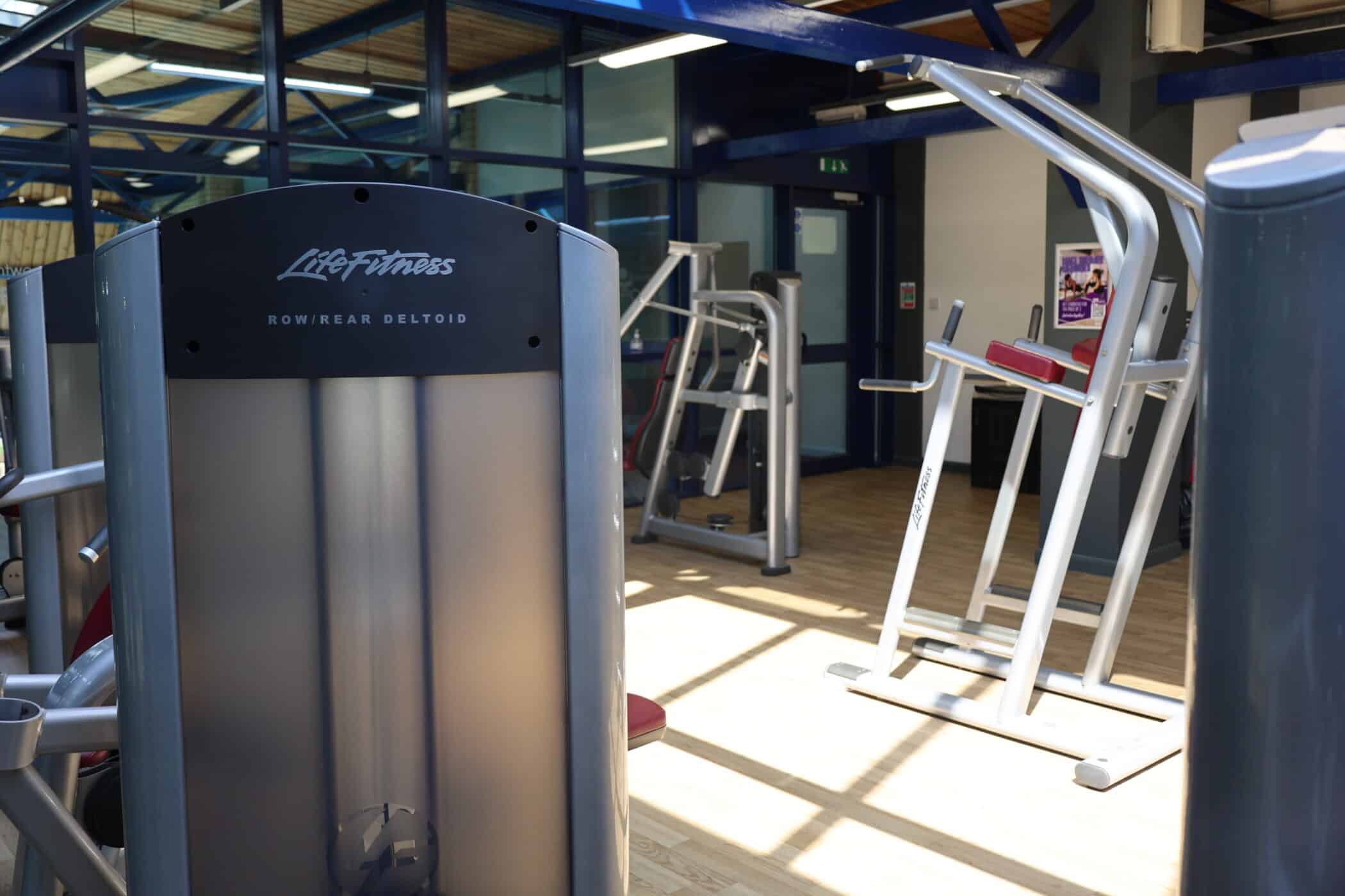 A gym interior at Brentwood School Sports Centre features various Life Fitness exercise machines, including a row/rear deltoid machine in the foreground, on a wooden floor with natural light streaming through large windows.