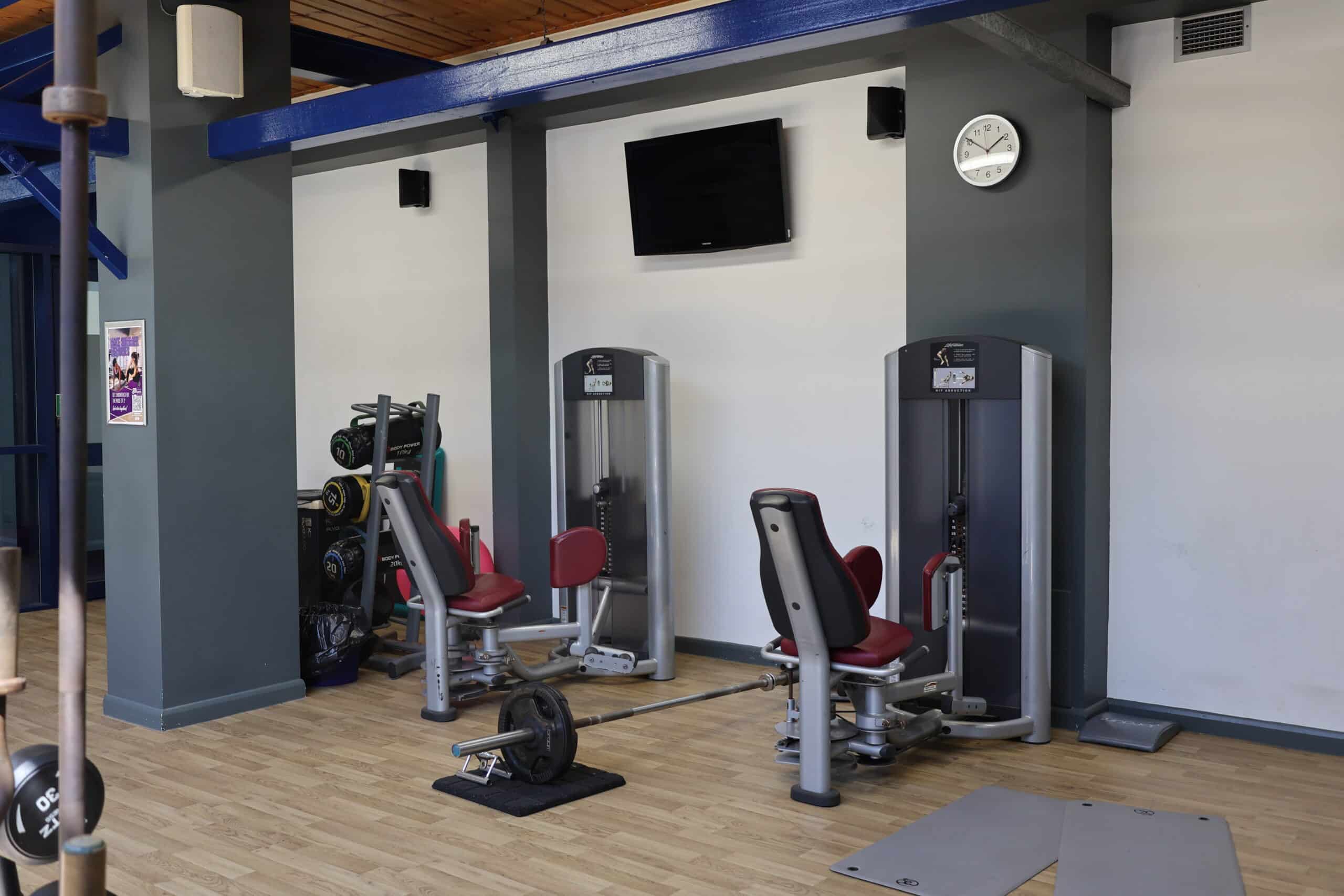 A gym with two weight machines, a barbell on the floor, a rack of weights, wall-mounted TV, clock, and light wood flooring. The walls are grey and white with blue ceiling beams. Sports Centre Online Booking is available for easy access.
