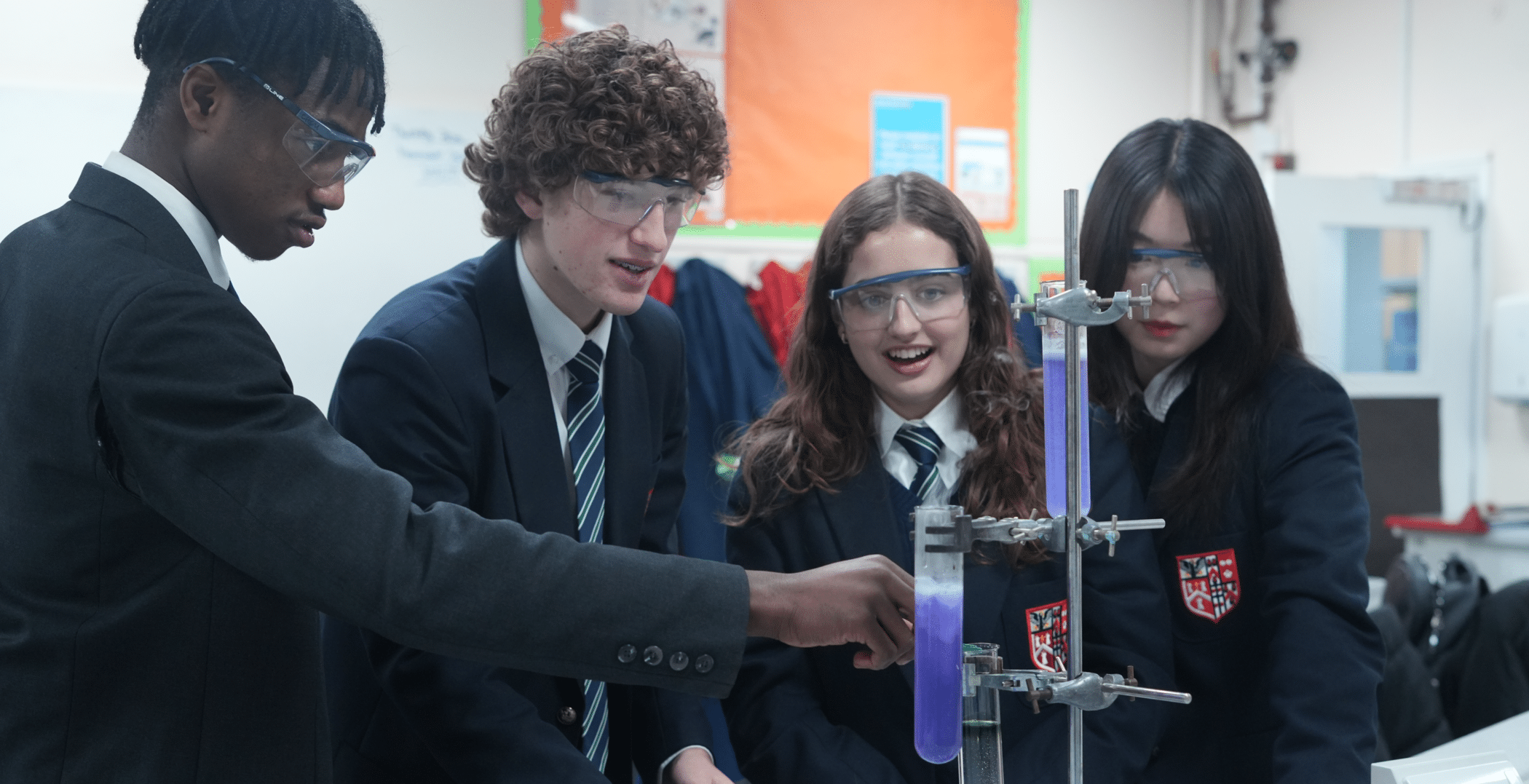 Four students in school uniforms and safety goggles conduct a science experiment in a classroom, exploring Sixth Form Academic Pathways as they observe a chemical reaction with purple liquid in a glass tube apparatus. They appear engaged and focused.