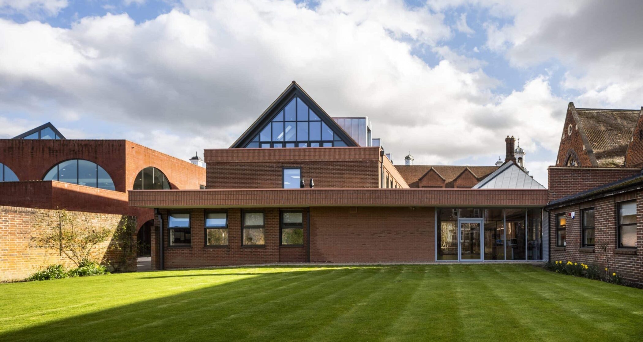 A modern brick building with large triangular glass windows and an arched walkway, surrounded by neatly trimmed grass under a partly cloudy sky, offers striking design.