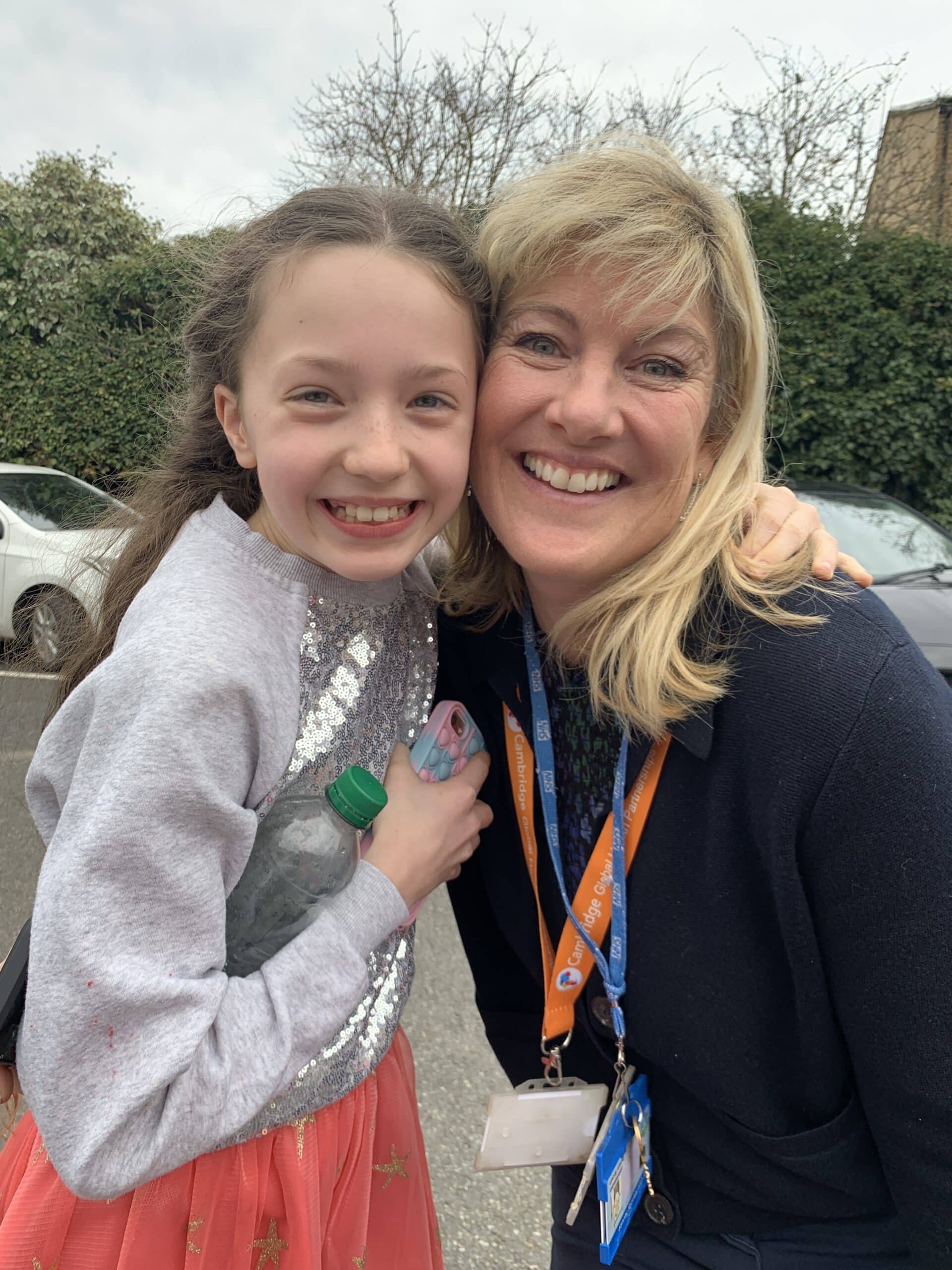 A young girl in a pink skirt hugs an older woman with blonde hair. They are both smiling at the camera, standing outdoors in a car park with trees and cars in the background.