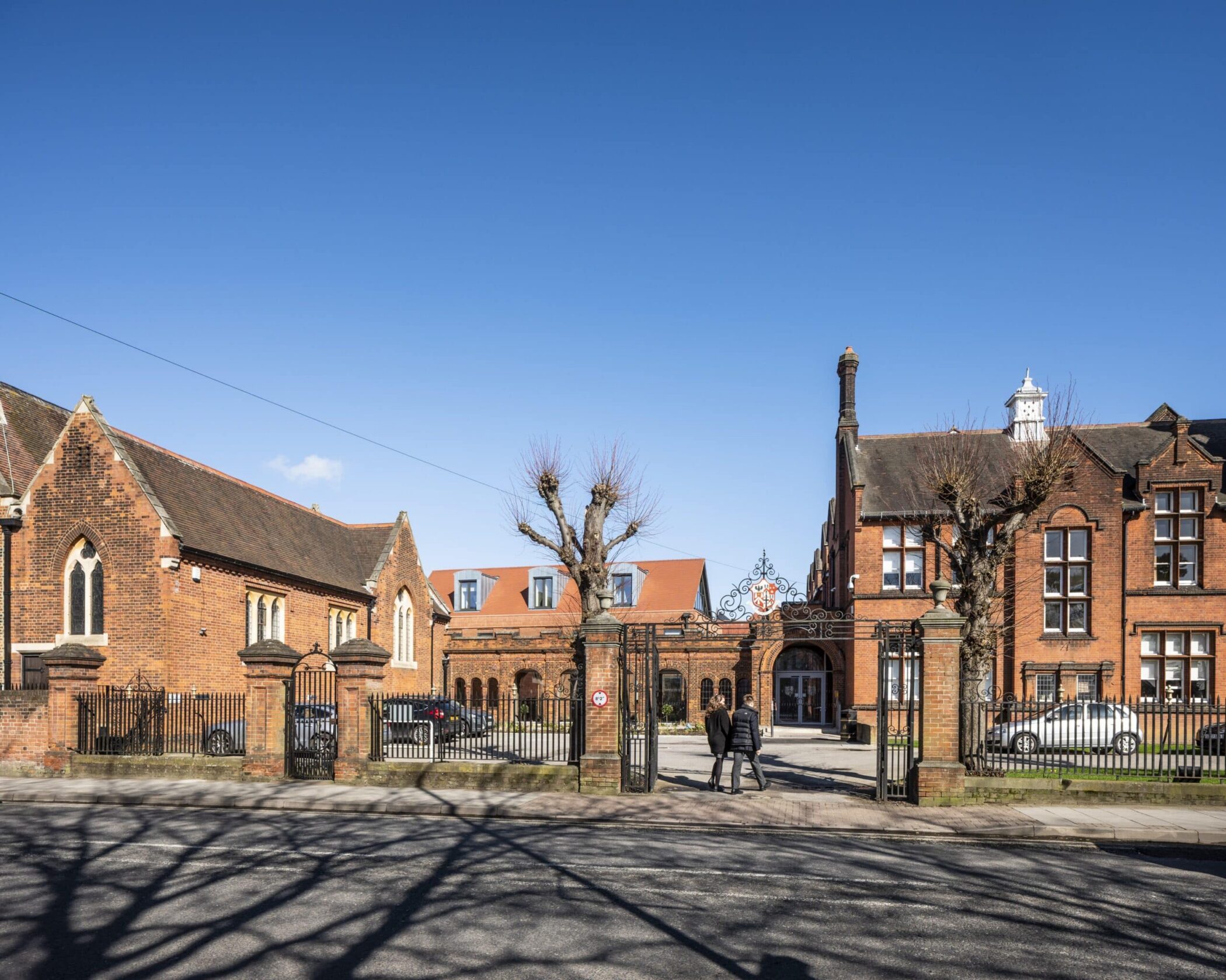 Two people walk towards the entrance of a large, historic red-brick building complex with iron gates, leafless trees, and parked cars, under a clear blue sky.