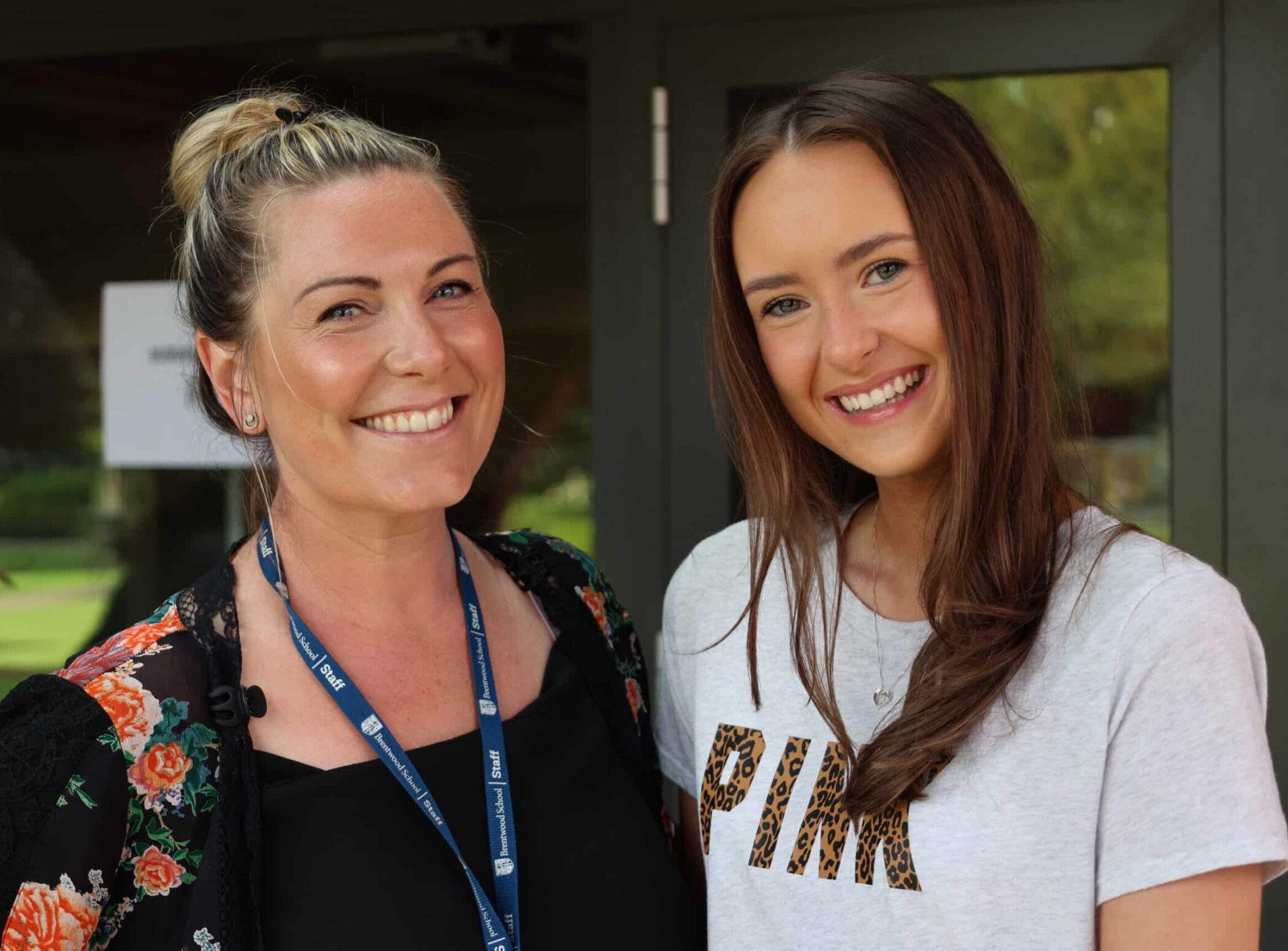 Two women smiling whilst standing close together outdoors, one wearing a lanyard and floral-patterned jacket, the other in a white T-shirt with PINK written on it in animal print letters.