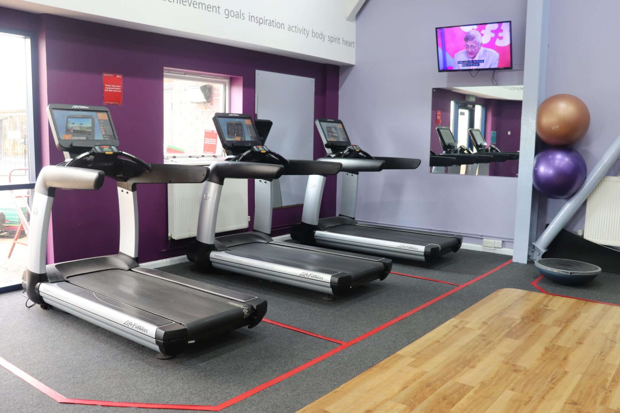 Three running machines are lined up in a gym with purple walls. Two large exercise balls and a balance trainer are in the corner, near the TV screen mounted above. Check Sports Centre Opening Times to plan your next workout session.