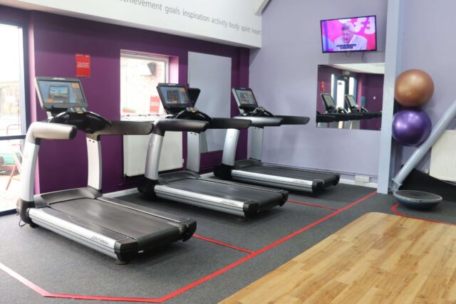 Three running machines are lined up in a gym with purple walls. Two large exercise balls and a balance trainer are in the corner, near the TV screen mounted above. Check Sports Centre Opening Times to plan your next workout session.