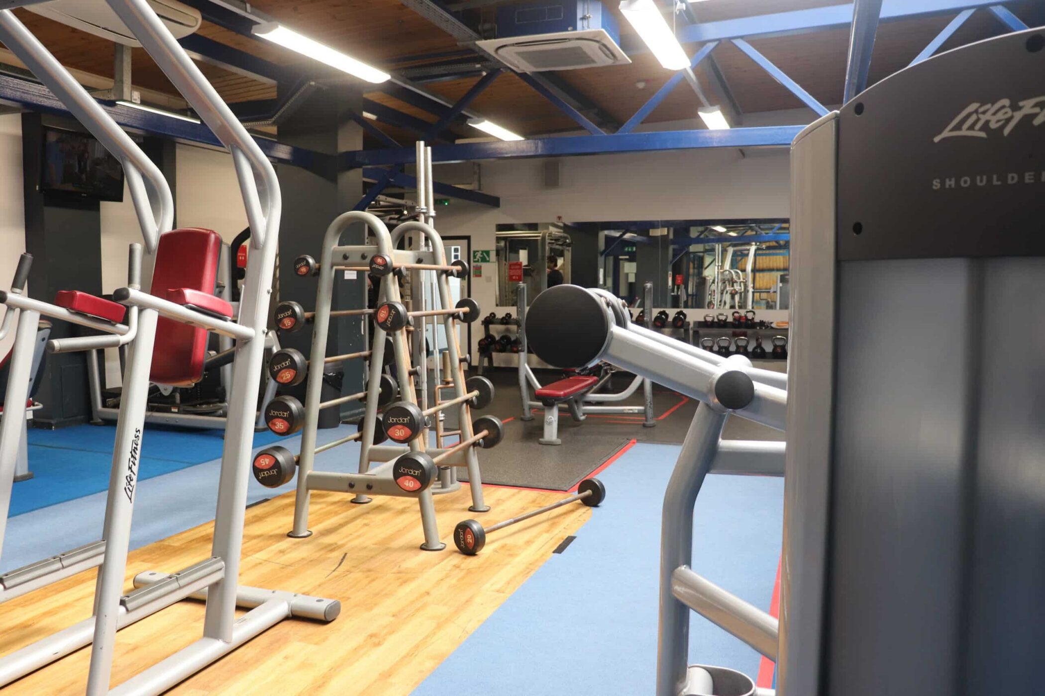 A modern gym with diverse equipment—weight racks, resistance machines, benches, and mirrors—in a brightly lit room with blue beams and wooden ceiling accents. Check Sports Centre Opening Times to plan your next workout.