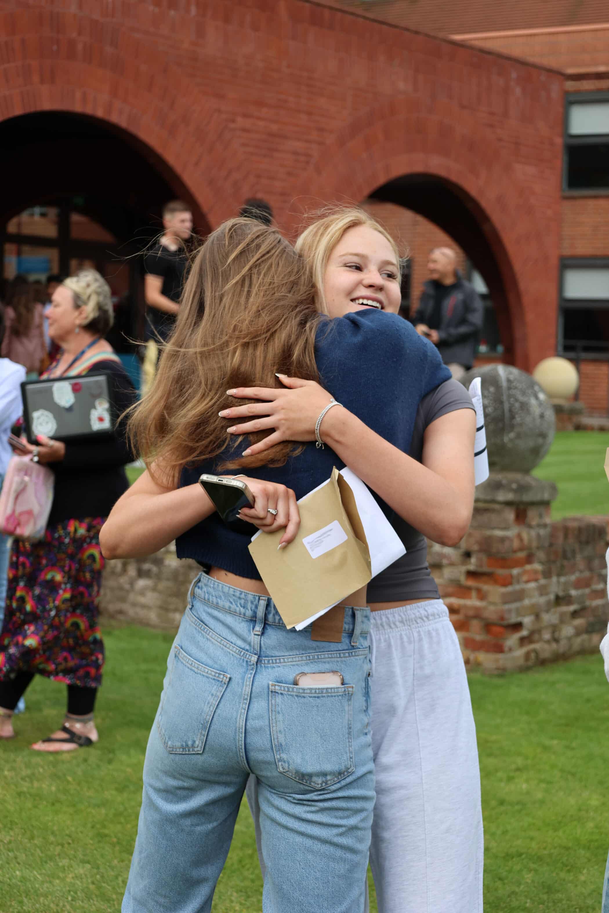 Two young women hug outdoors, smiling happily. One holds a brown envelope, suggesting a celebration or results day. People and a red-brick building are visible in the background.