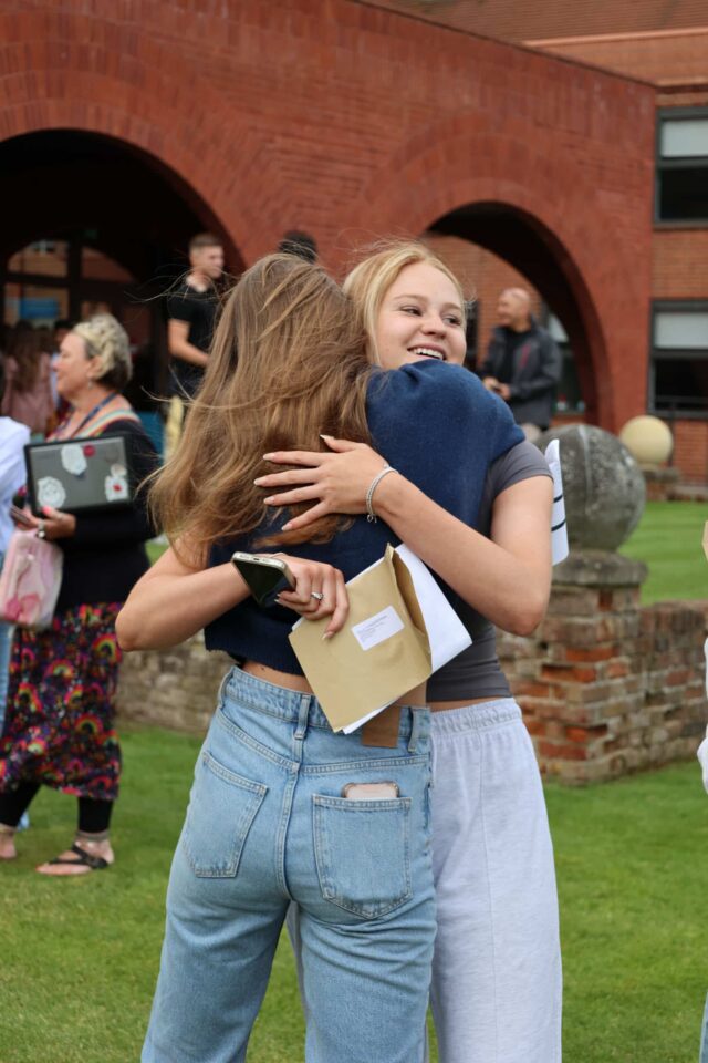 Two young women hug outdoors, smiling happily. One holds a brown envelope, suggesting a celebration or results day. People and a red-brick building are visible in the background.