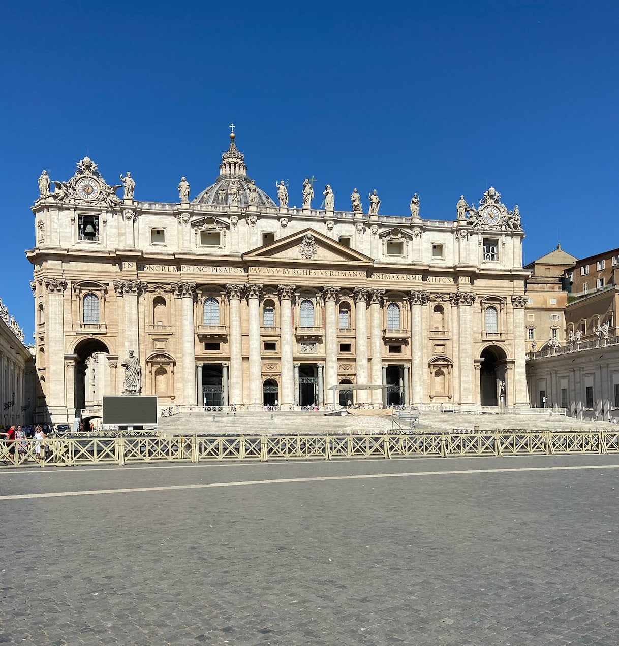 St Peter’s Basilica in Vatican City, seen from St Peter’s Square under a clear blue sky, with its grand façade, columns, statues, and a central dome visible above the entrance.