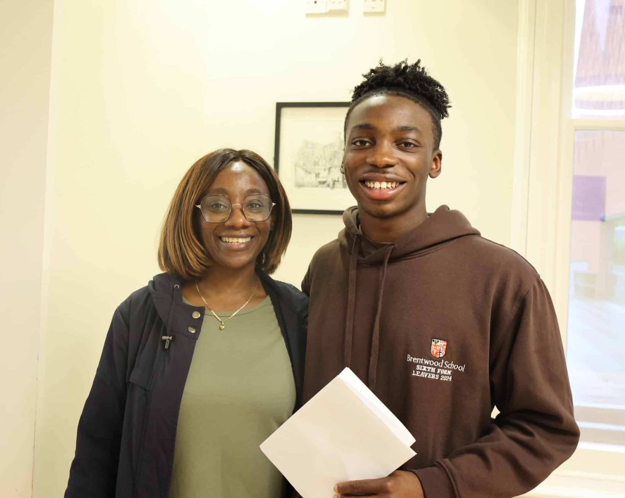 A smiling young man holding papers stands next to a smiling woman in glasses. He wears a brown hoodie with a school logo; she wears a green top and dark jacket. They are indoors with a framed picture on the wall behind them.
